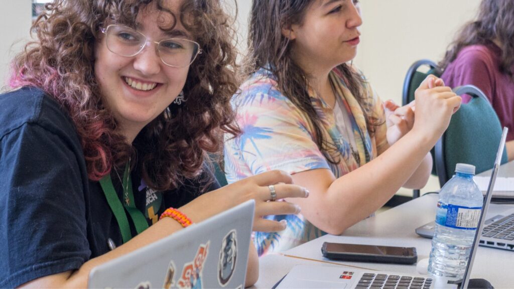 A row of young women sit at a classroom table with laptops, phones, and a water bottle in front of them