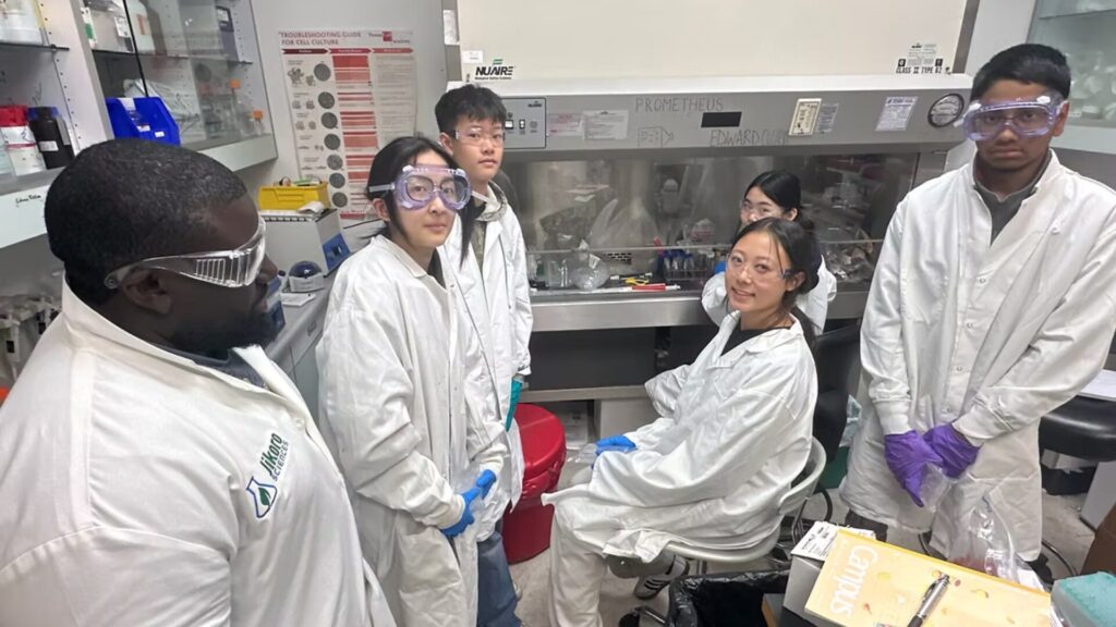 A researcher in a lab coat and safety goggles instructs a group of high school students, also wearing lab coats and goggles, gathered around a biosafety cabinet during a hands-on biomedical research session.