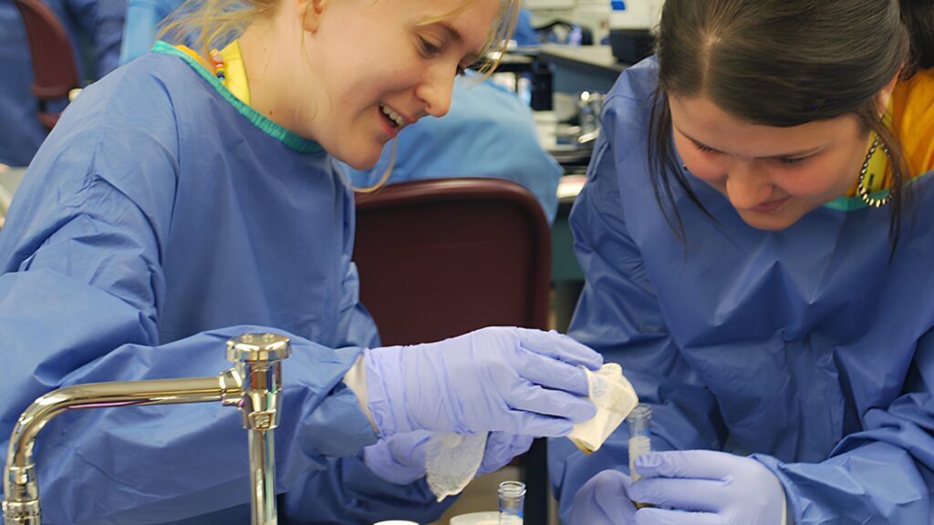 Two students wearing blue gowns and gloves and performing an experiment