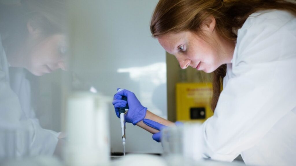 A student crouching over a table, wearing a lab coat and purple gloves while holding a syringe-esque structure in one hand