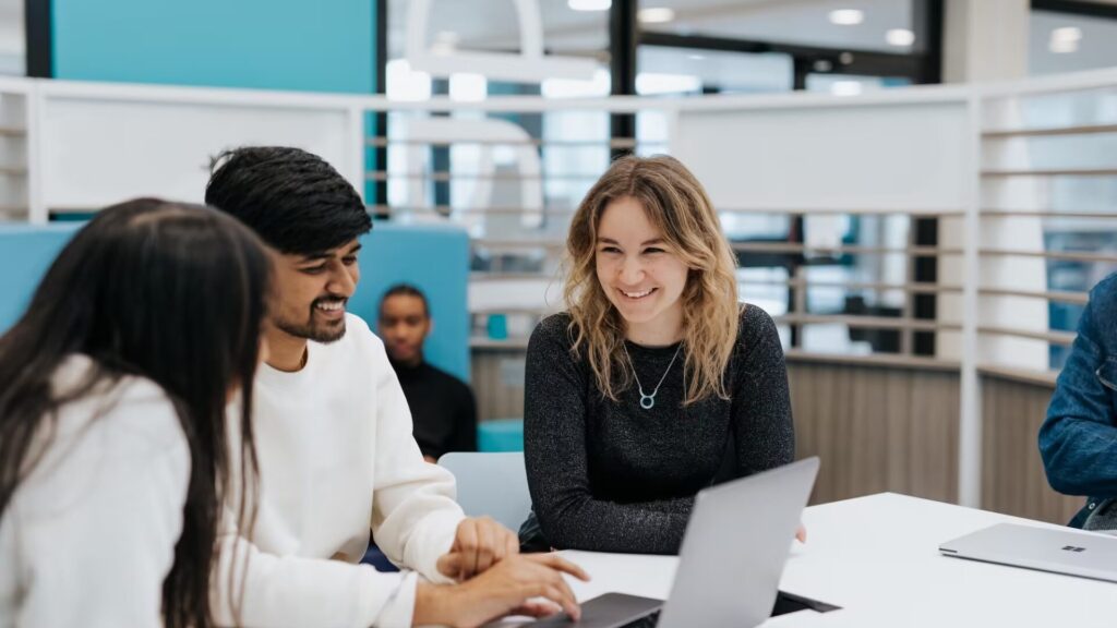 A trio of students sitting around a table, talking to each other, with one of them working on a laptop