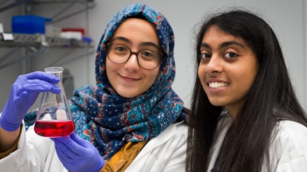 Two students looking into the camera, with one of them holding a beaker of red liquid, and smiling