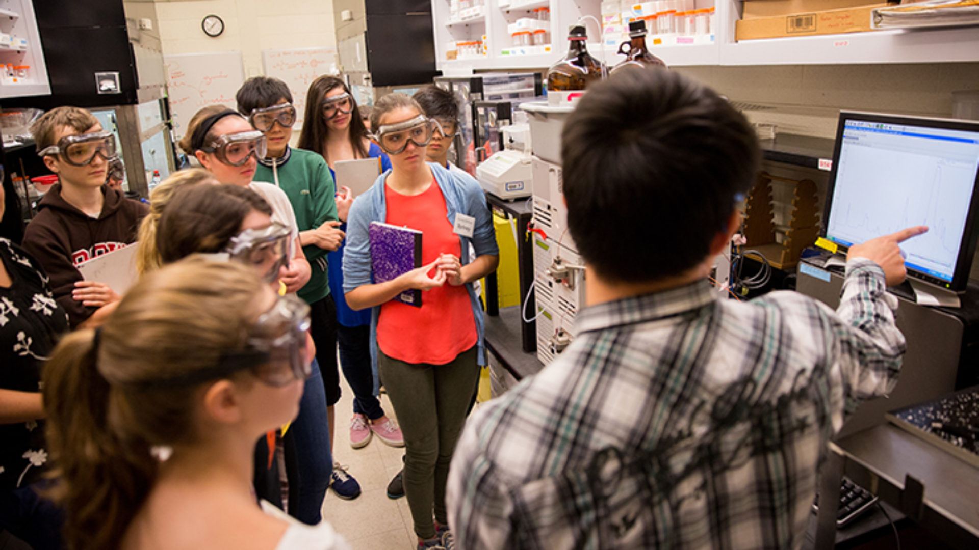 A lab instructor points to a display where multiple high school students wearing safety goggles gather around to listen.