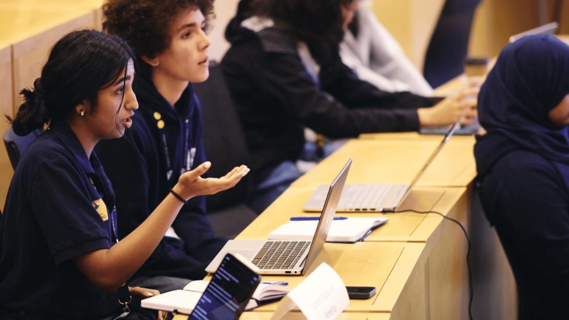A classroom, focusing on a row of high school students who have devices and notebooks on their tables; a girl, seated, is speaking and gesturing.