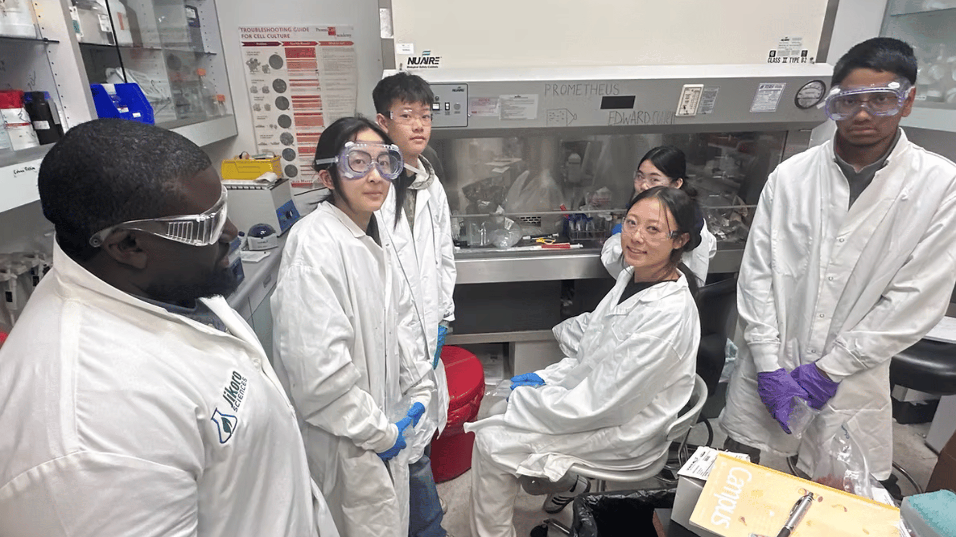 A researcher in a lab coat and safety goggles instructs a group of high school students, also wearing lab coats and goggles, gathered around a biosafety cabinet during a hands-on biomedical research session.
