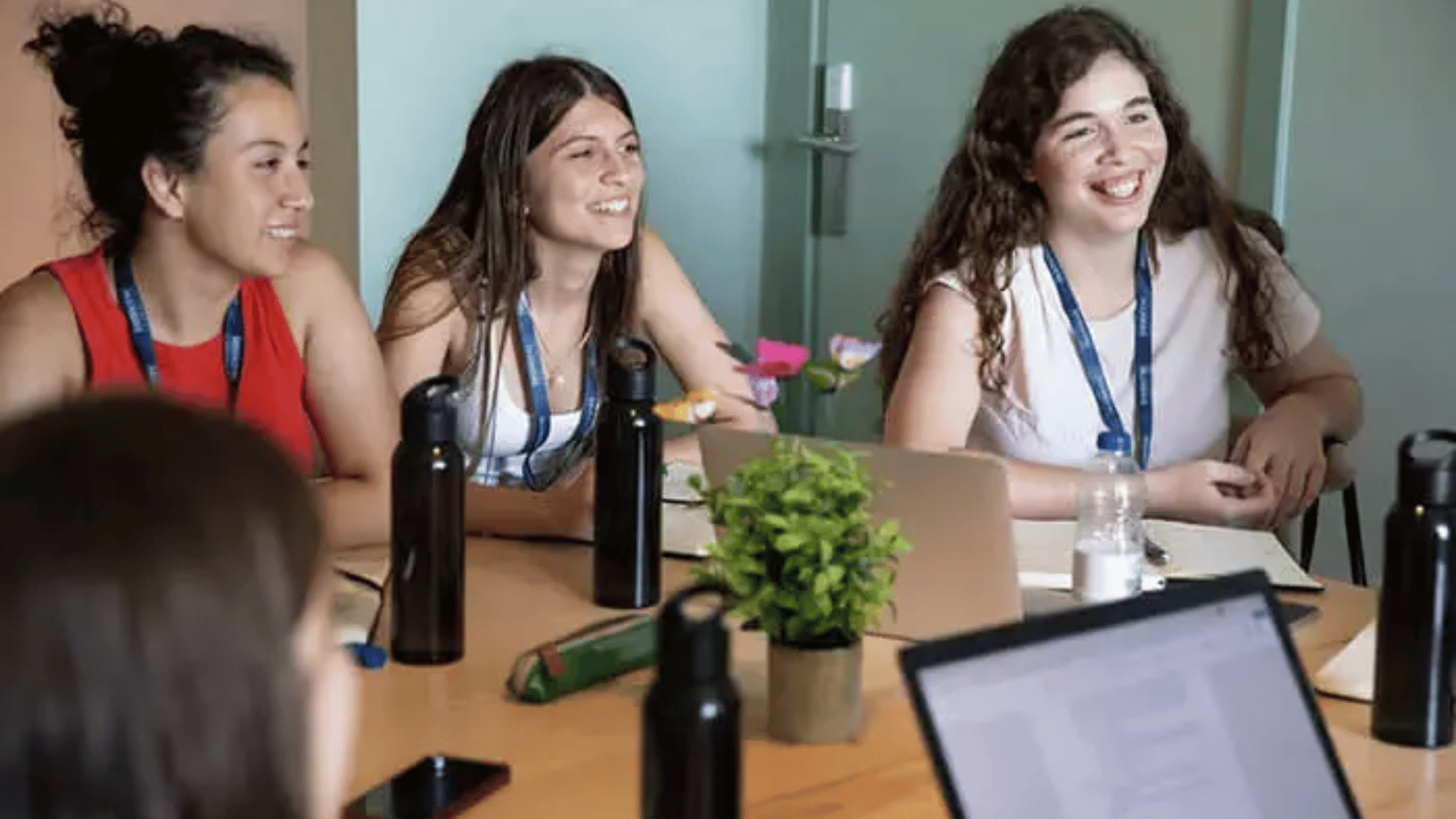 A group of high school students sitting around a table with laptops and water bottles on it, listening to someone speaking.