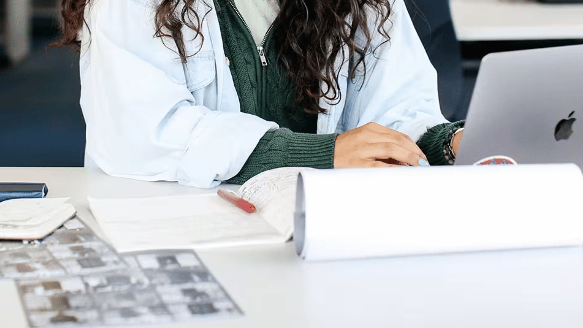 A student sitting next to a table, typing something on a grey macbook, with a notebook in front of them