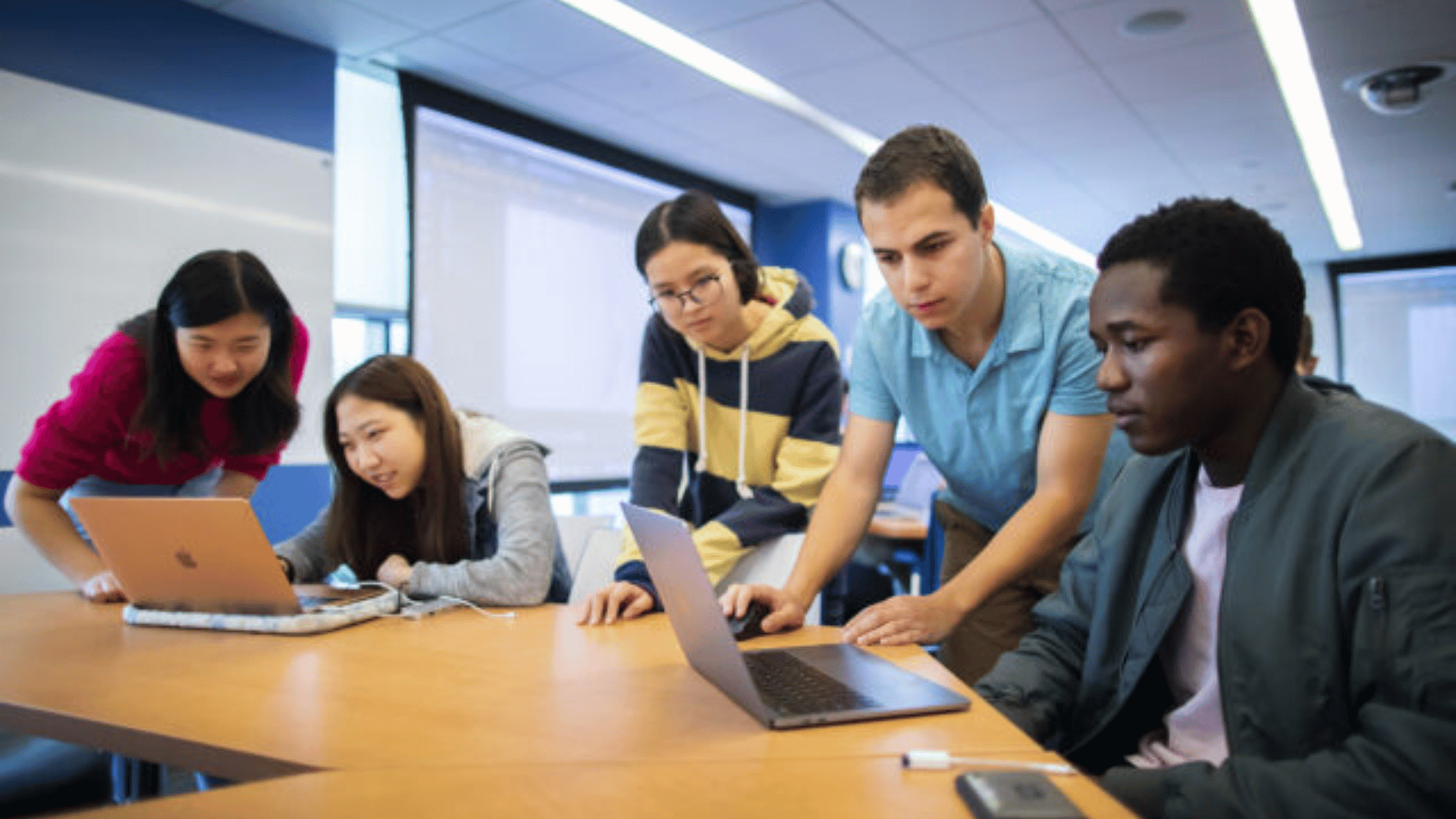 Five students sitting around a table together and working on something across two laptops