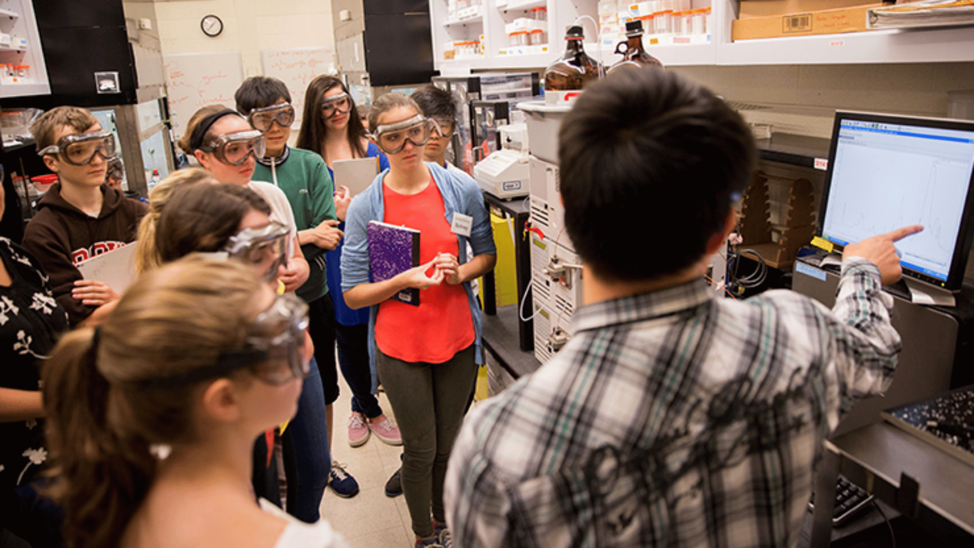A lab instructor points to a display where multiple high school students wearing safety goggles gather around to listen.