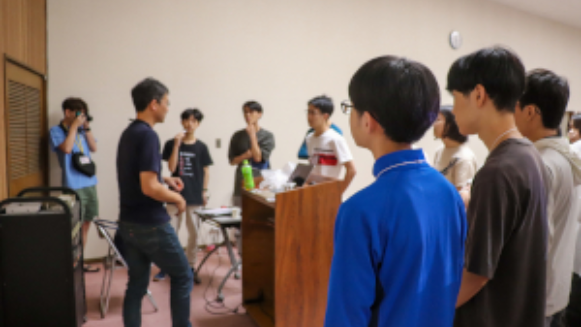 A group of students standing around a podium, behind which an instructor is standing and explaining something