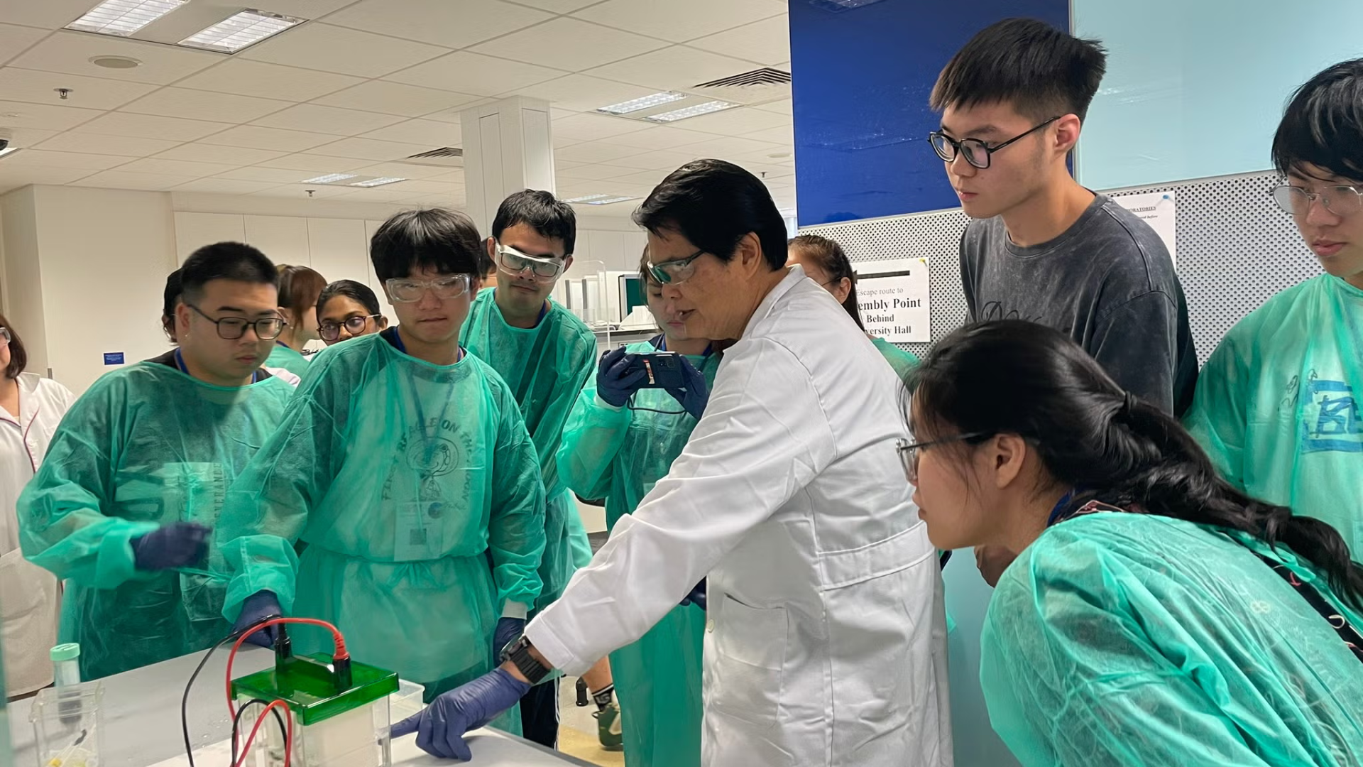 A group of students in lab uniforms look on as a teacher explains a concept to them in a lab.