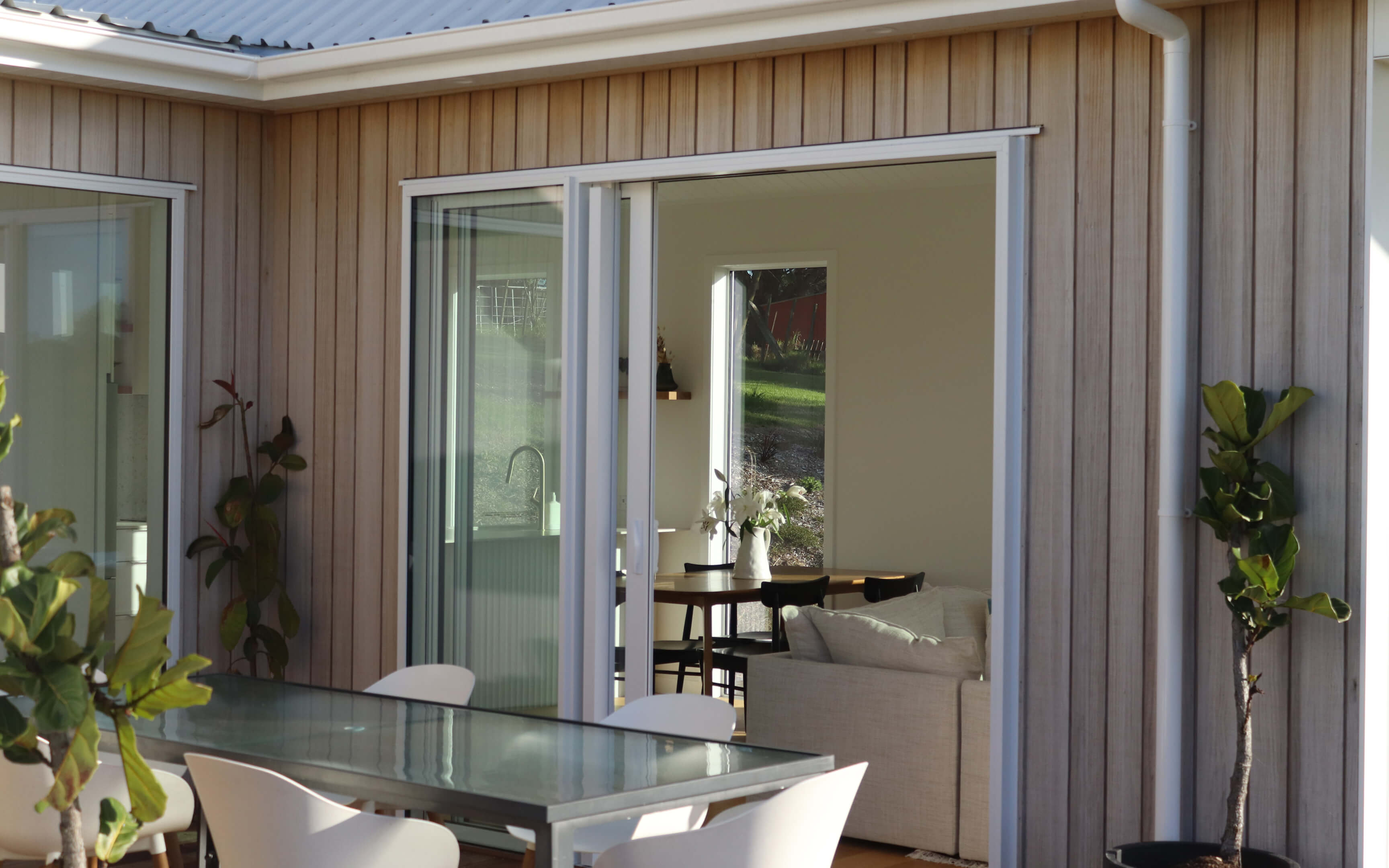 View through a sliding glass door into a bright, modern living and dining area, with a glass outdoor table and white chairs on a timber deck surrounded by vertical wood cladding.