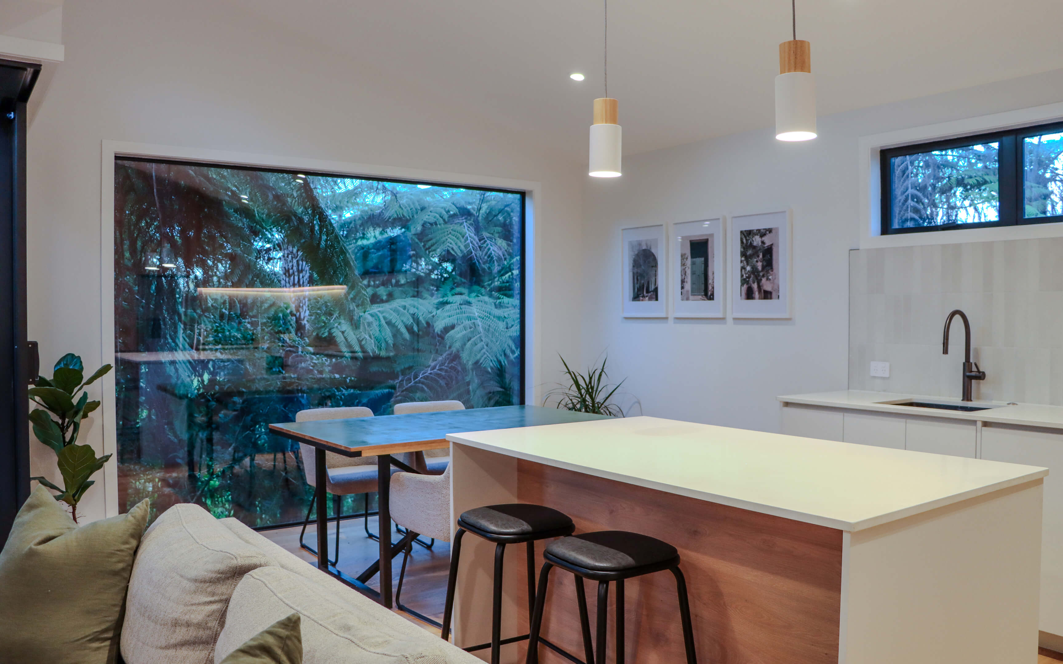 Modern open-plan kitchen and dining area with a large window overlooking lush native bush, featuring a central island with bar stools, pendant lighting, and minimalistic decor.