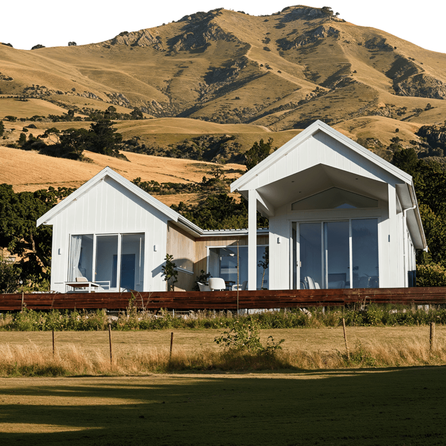 Stylish white minor dwelling with two gabled roofs and full-height windows, set on a timber deck with golden hills rising in the background.