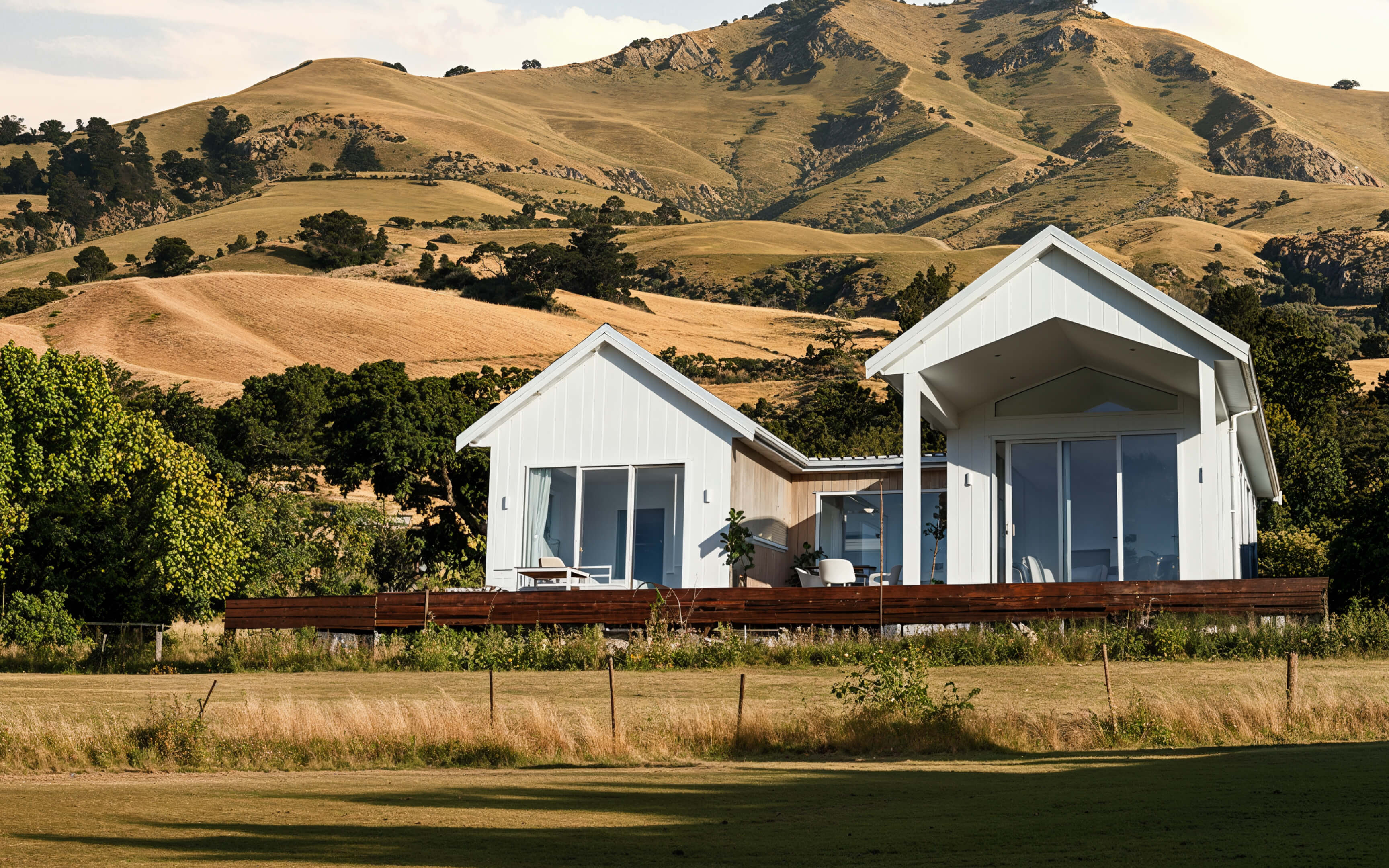 Modern white minor dwelling with twin gabled roofs and large sliding glass doors, positioned on a wooden deck and surrounded by grassy fields and rolling golden hills.