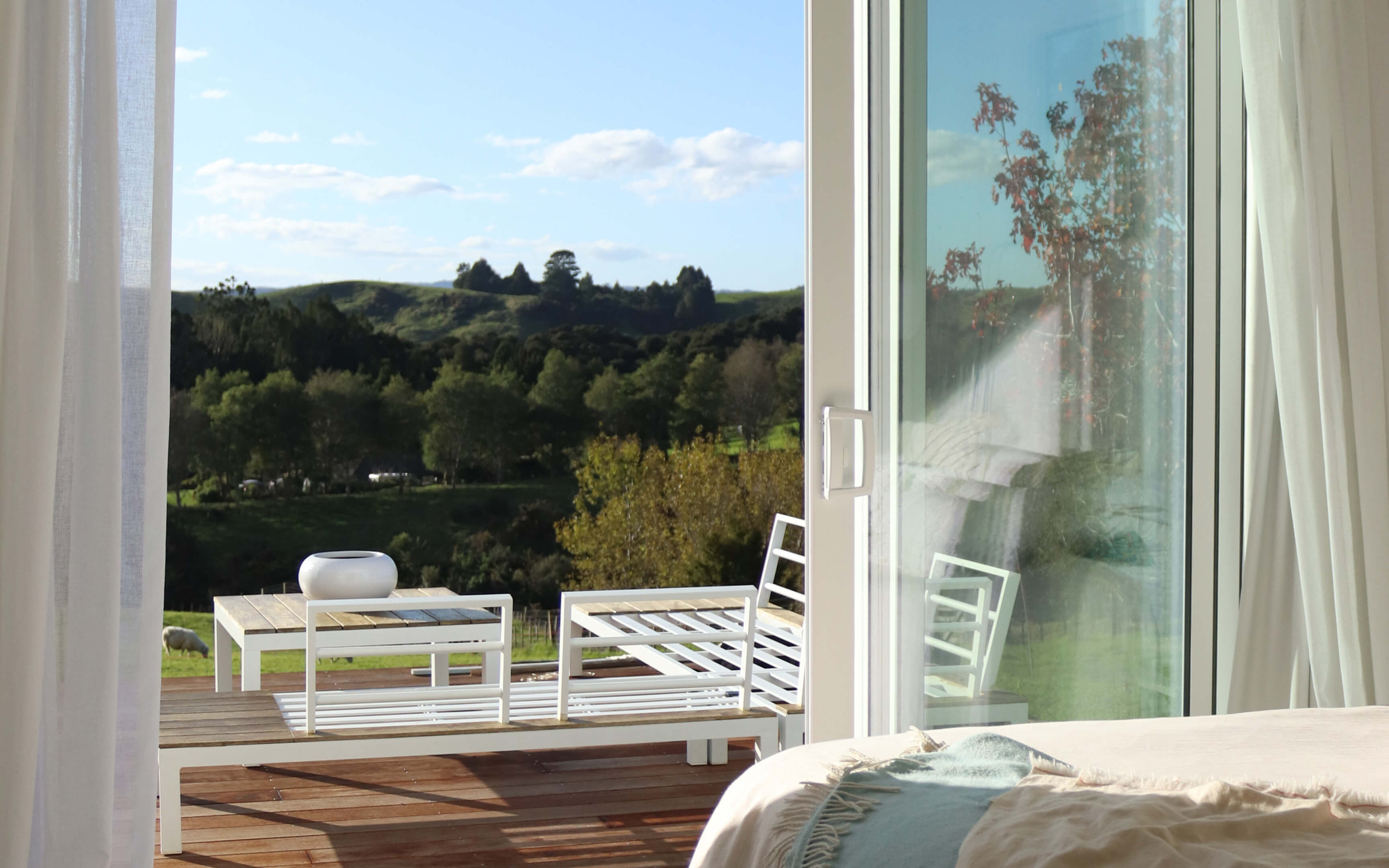 View from a bedroom through sliding glass doors onto a timber deck with white outdoor furniture, overlooking green hills, trees, and grazing sheep under a blue sky.