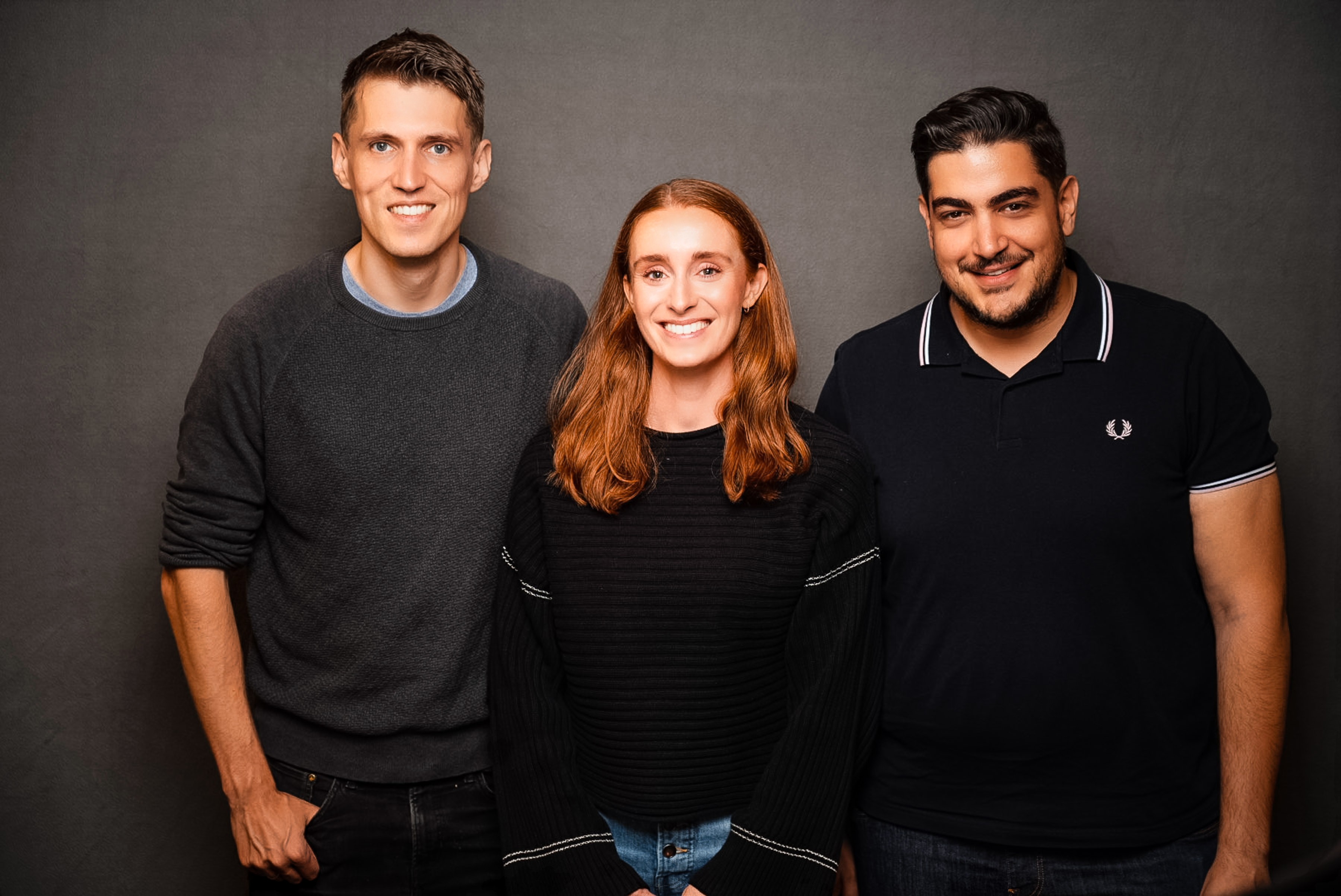Three smiling adults standing shoulder to shoulder against a dark gray background, a man in a dark sweater on the left, a woman with red hair in a black sweater in the center, and a man in a black polo shirt on the right.