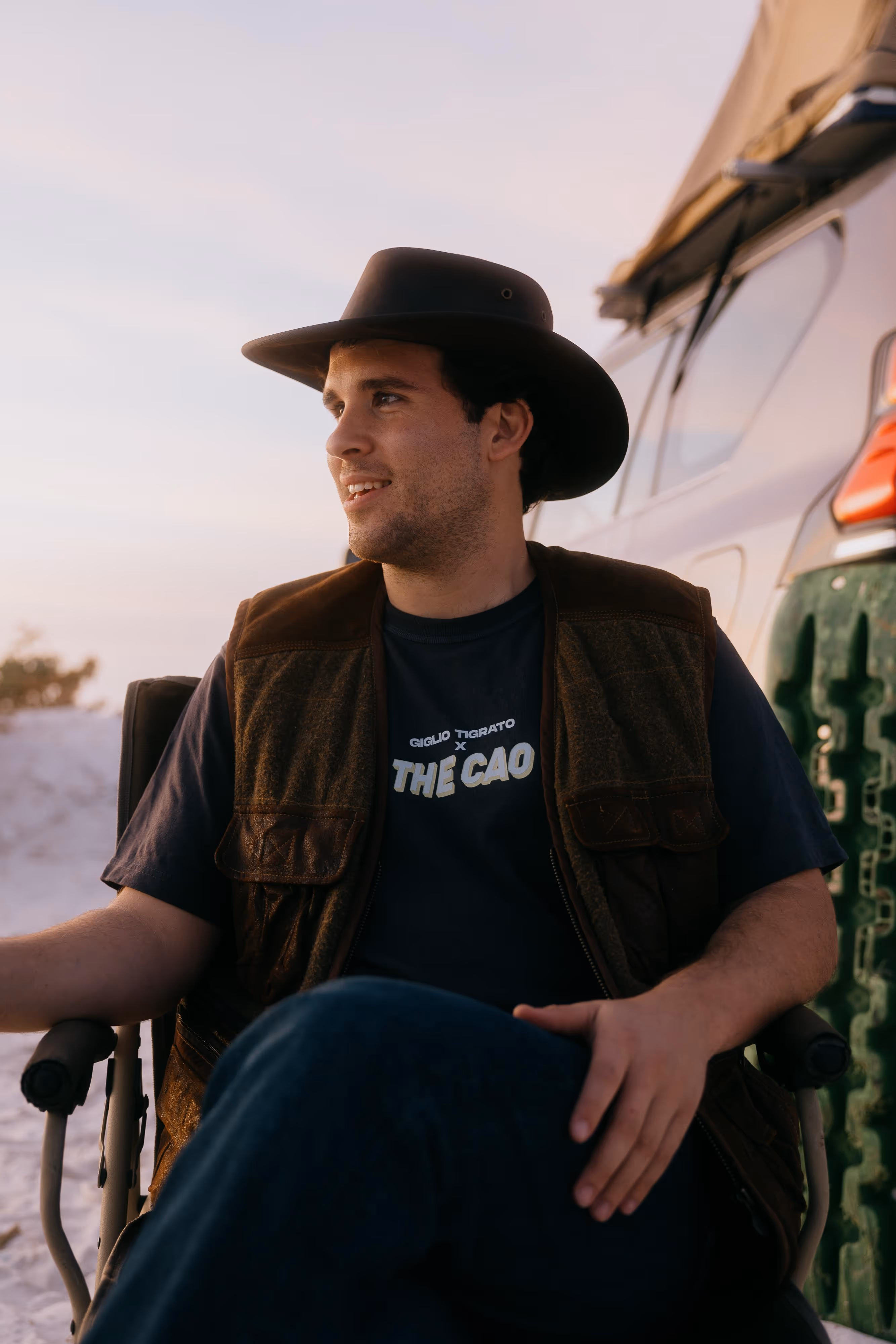 Man wearing a brown vest and black wide-brim hat sitting in a chair next to a vehicle during sunset.
