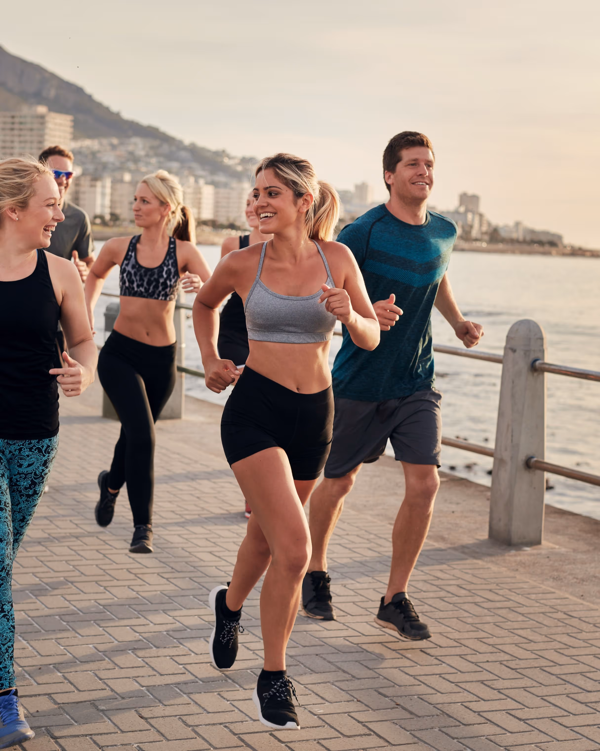 Group of diverse young adults jogging along a waterfront promenade in athletic wear.