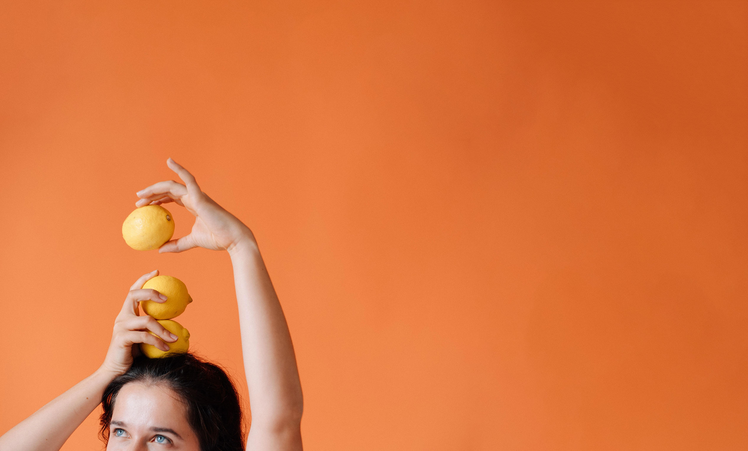 Eine Frau mit dunklen Haaren und blauen Augen balanciert drei Zitronen auf ihrem Kopf. Mit der einen Hand hält sie zwei Zitronen. Mit der anderen Hand hält sie eine Zitrone. Von der Frau sind nur die obere Hälfte des Gesichts zu sehen. Sie blickt nach links oben.
