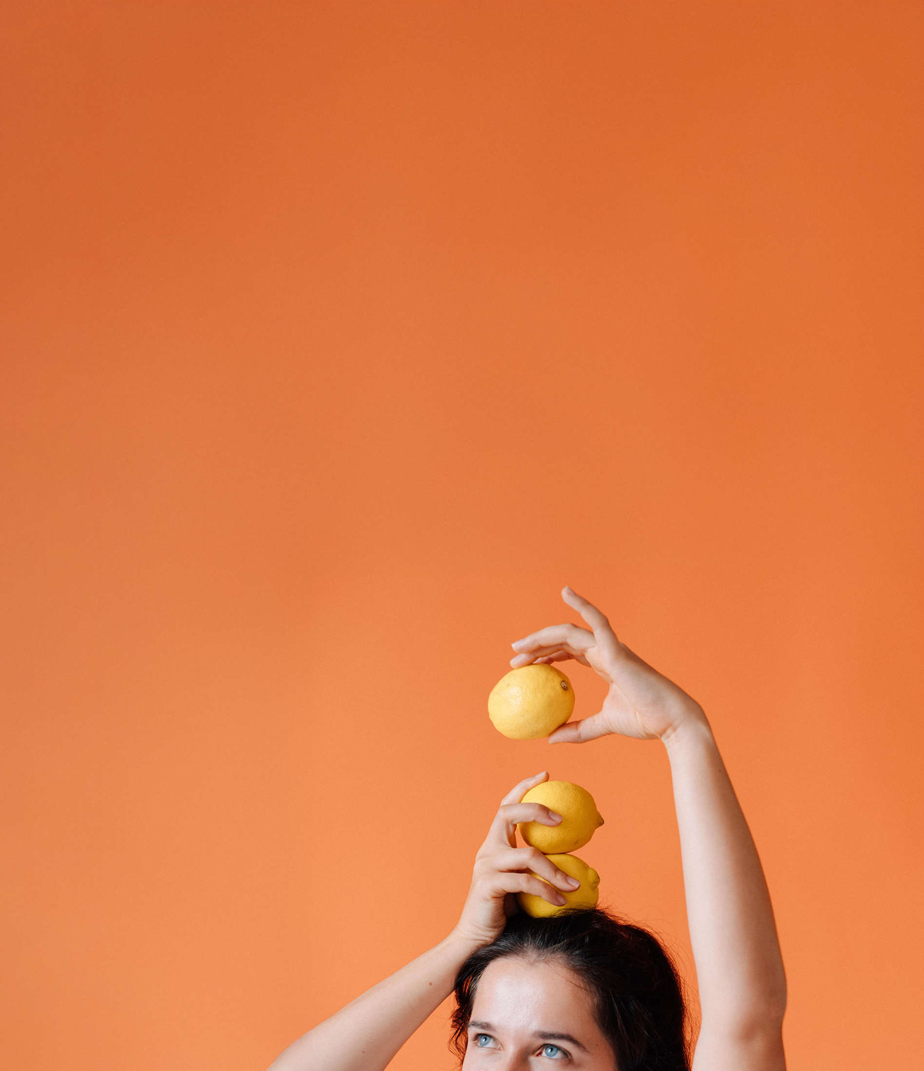Eine Frau mit dunklen Haaren und blauen Augen balanciert drei Zitronen auf ihrem Kopf. Mit der einen Hand hält sie zwei Zitronen. Mit der anderen Hand hält sie eine Zitrone. Von der Frau sind nur die obere Hälfte des Gesichts zu sehen. Sie blickt nach links oben.