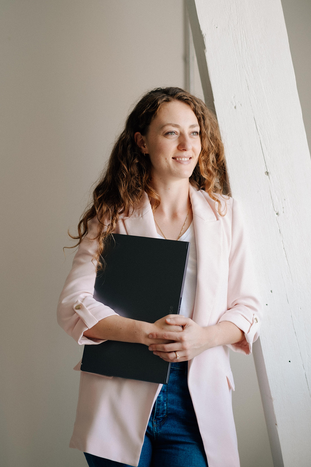 Eine Ernährungsberaterin ist an einen weissen Holzbalken gelehnt. Sie blickt lächelnd und selbstbewusst in die Ferne. Sie trägt einen hellem Blazer. In den Armen hält sie einen Laptop.