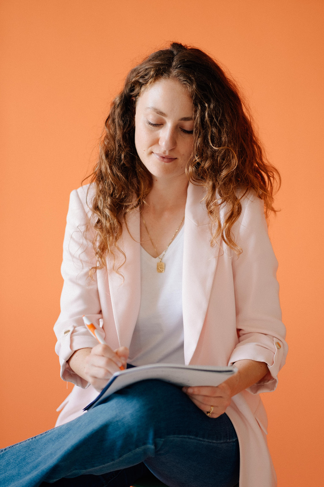Eine Frau sitzt vor einen orangen Hintergrund. Auf ihrem Schoss ist ein Schreibblock. Sie schreibt etwas mit einem Kugelschreiber.