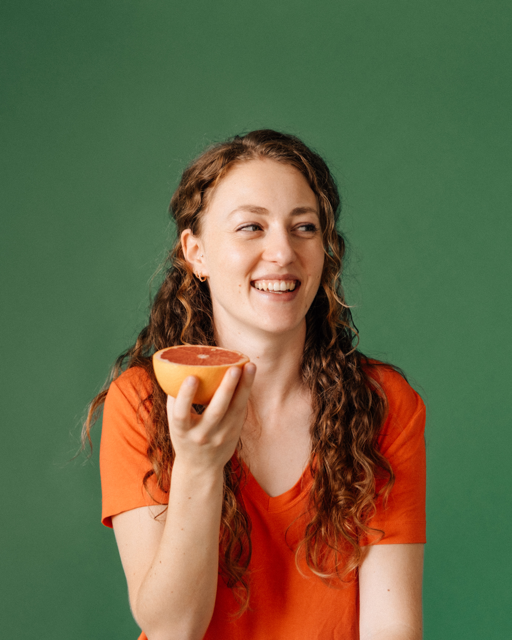 Eine Frau mit lockigem Haar hält lächelnd eine Hälfte einer Grapefruit in der Hand. Sie sitzt vor einer dunkelgrünen Wand und trägt ein oranges T-Shirt.