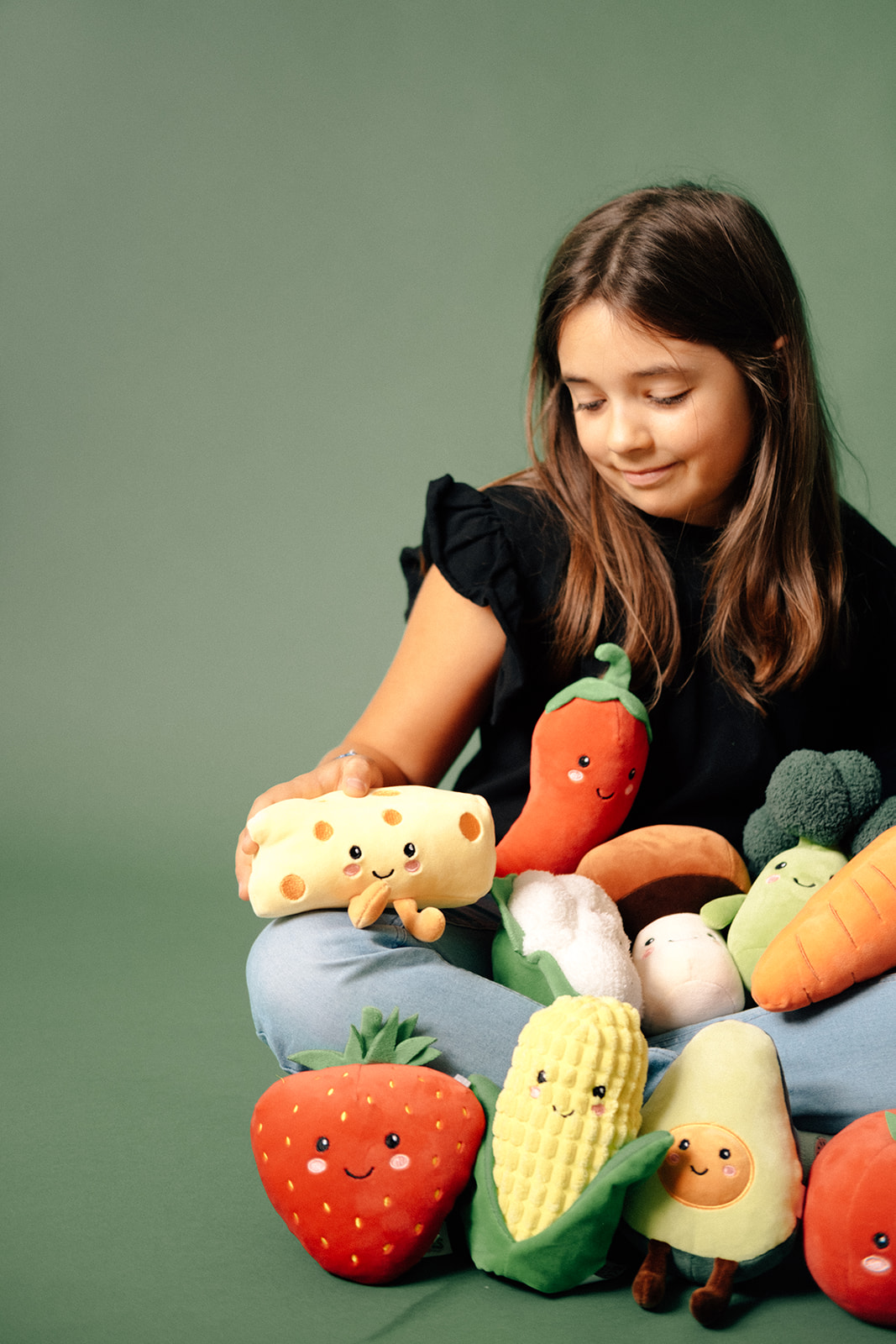 Ein Kind sitzt im Schneidersitz vor einer dunkelgrünen Wand. In ihrem Schoss und vor ihr befinden sich lauter verschiedene Lebensmittel aus Plüsch mit Gesichtern.