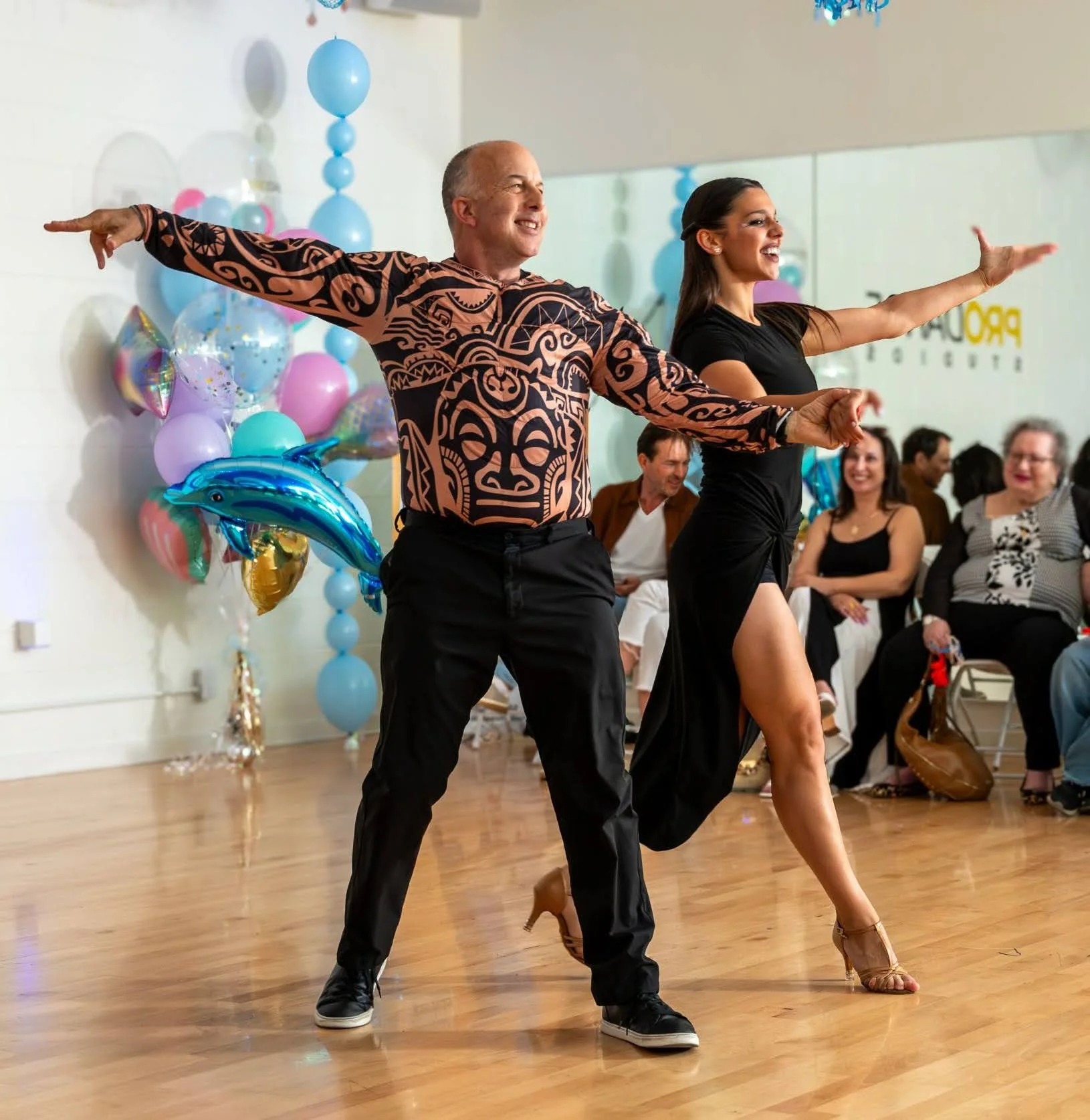 Social dance etiquette: a smiling dance couple strikes a pose with arms outstretched and hands joined on a studio floor, with balloons and seated spectators looking on at Pro Dance Studios.