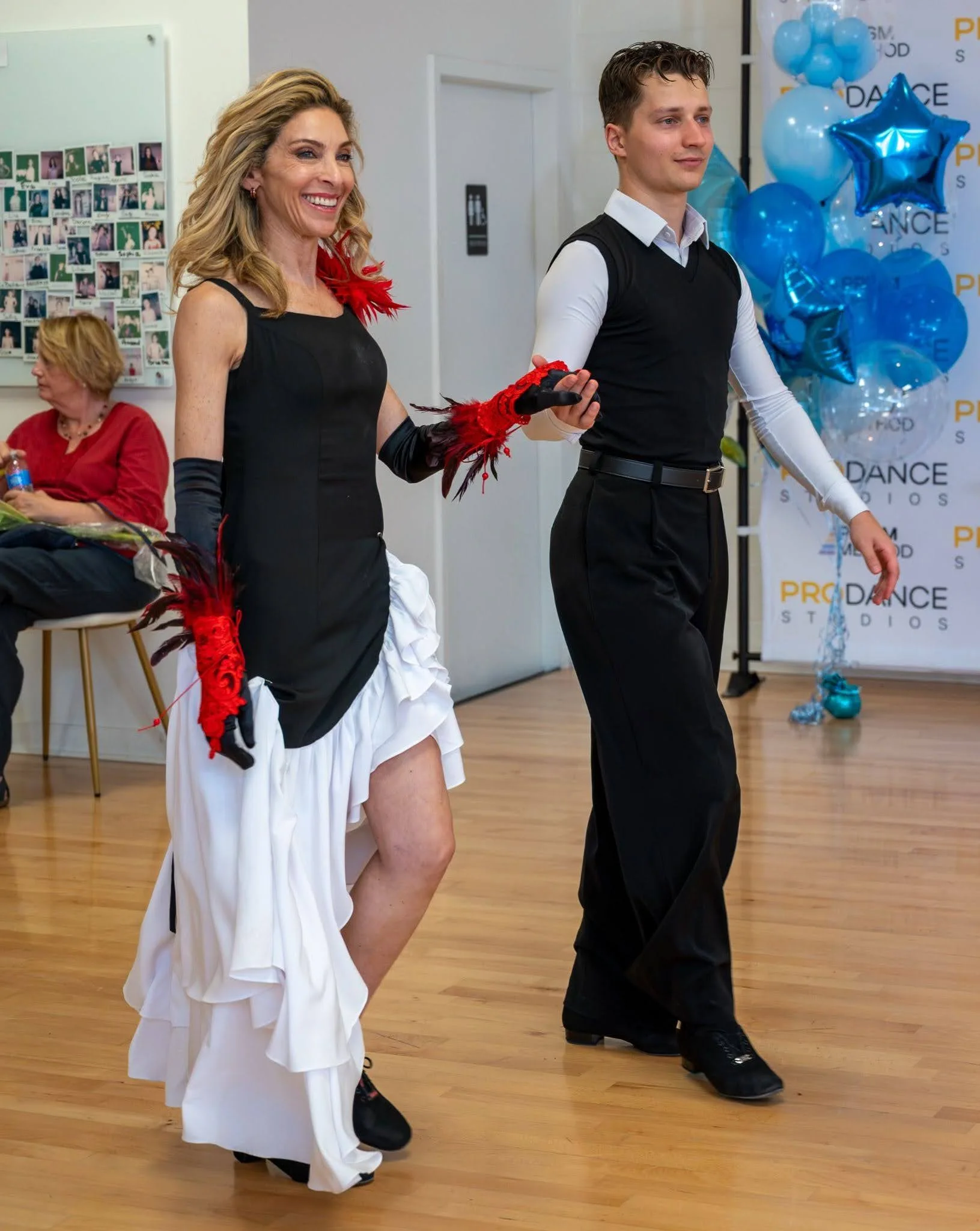 Social dance etiquette: a smiling woman in a black and white ruffled dress with red feathered gloves walks hand in hand with a man in a black vest on a studio dance floor.