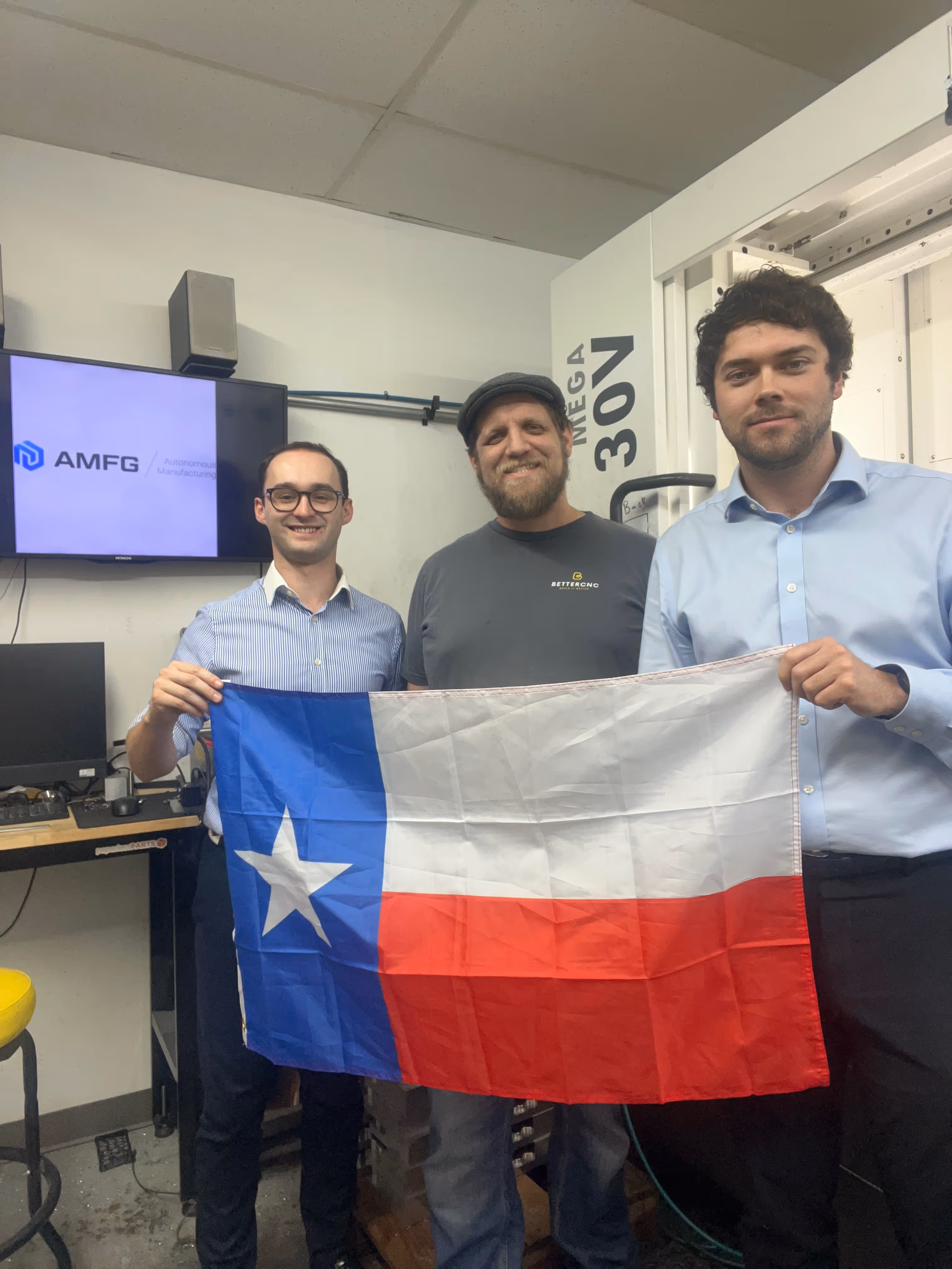 Jesse Casto, owner of BetterCNC, standing in his shop. He is with two AMFG employees, and all three of them are holding the Texas flag.