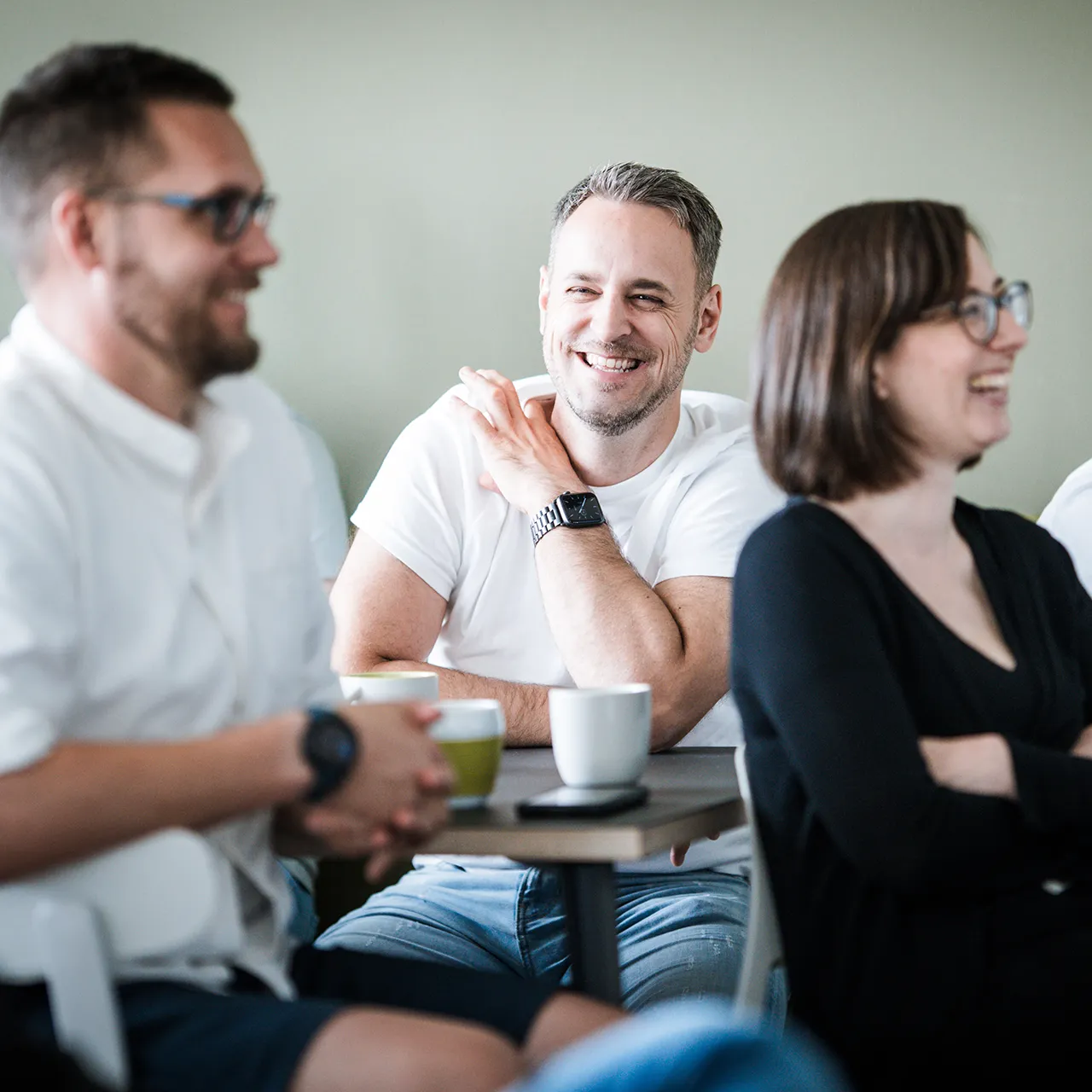 Three people sitting at a table, smiling and engaged in conversation, with mugs and a phone on the table.