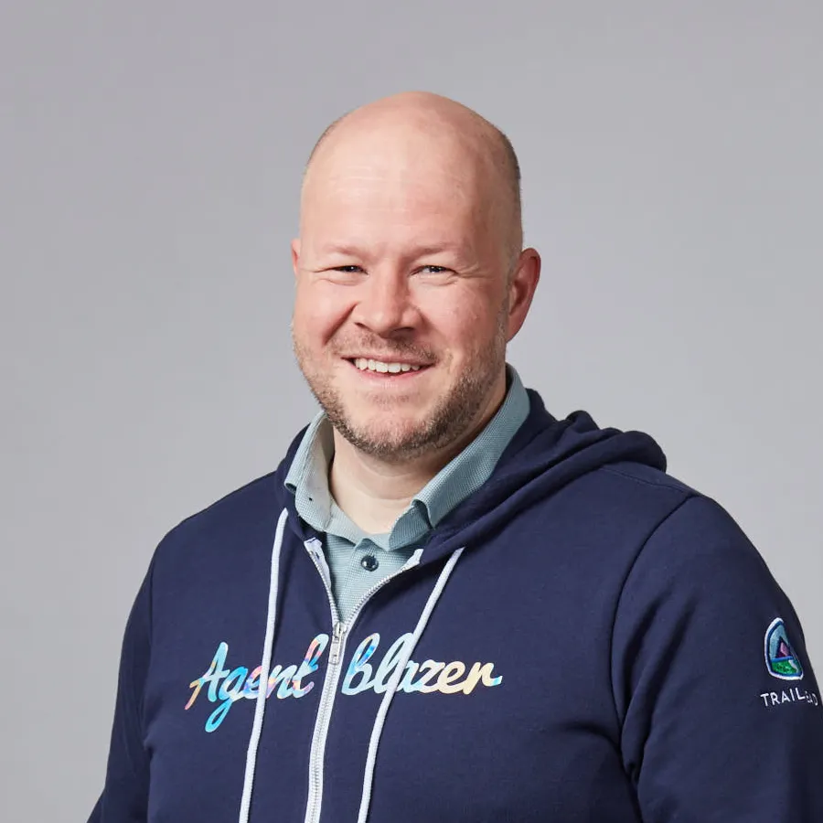 Smiling bald man wearing a blue hoodie with 'Agent Blazer' text and a logo patch on sleeve against a gray background.