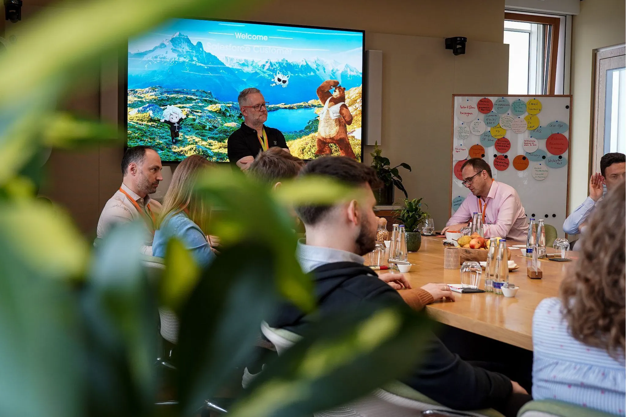 Group of professionals in a meeting room with a man standing near a screen displaying a nature-themed welcome slide and a whiteboard with colorful notes behind another man at the table.
