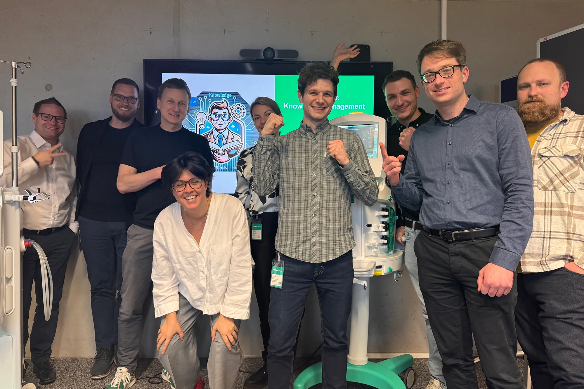 Group of nine smiling people posing indoors around medical equipment and a screen displaying a knowledge-themed graphic.