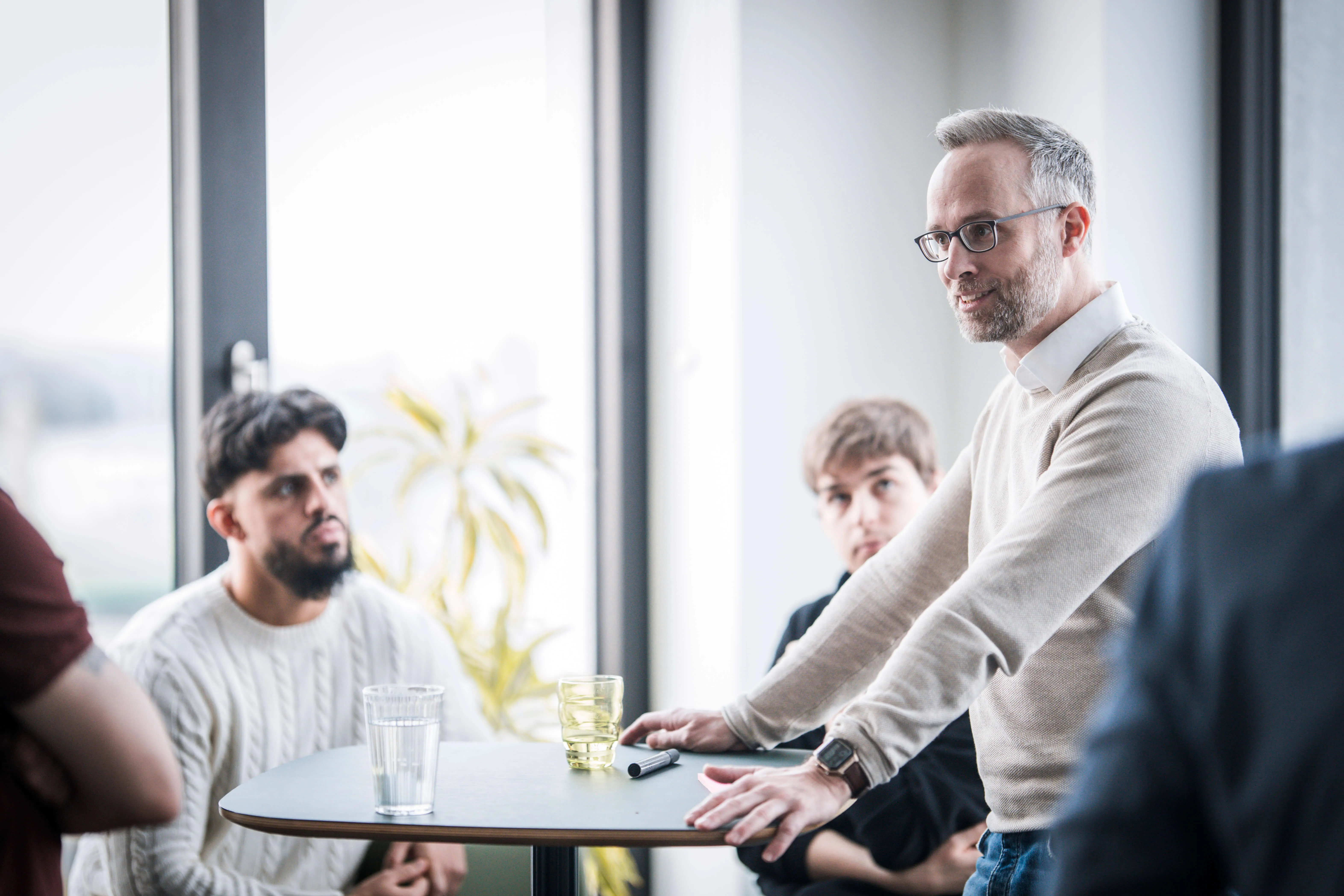 Man with glasses and beige sweater speaking to attentive group around a round table with glasses and marker.