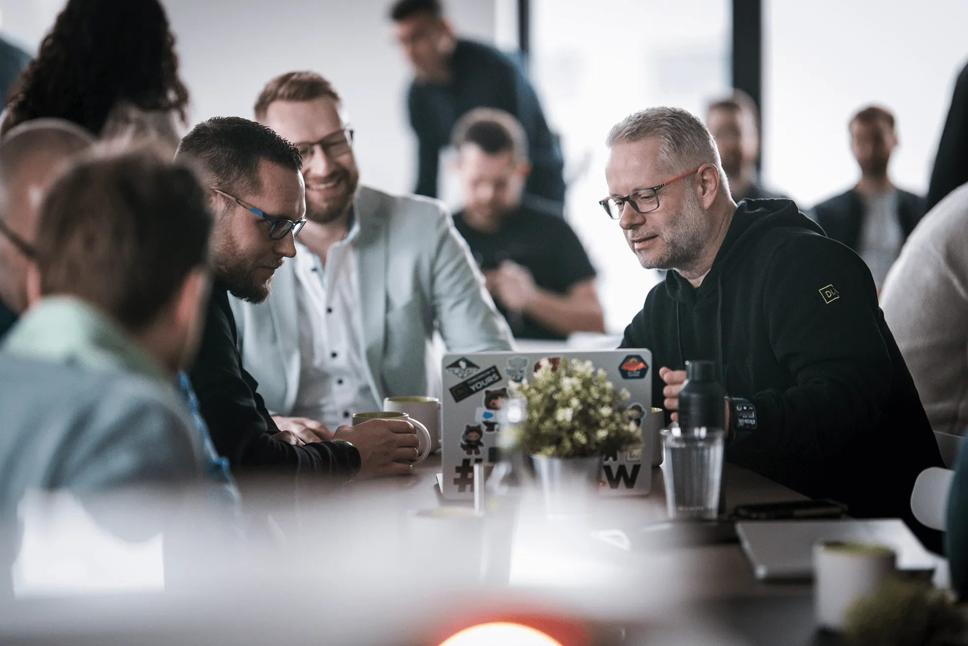 Group of people in a casual meeting around a table with a laptop and coffee mugs in a bright office.