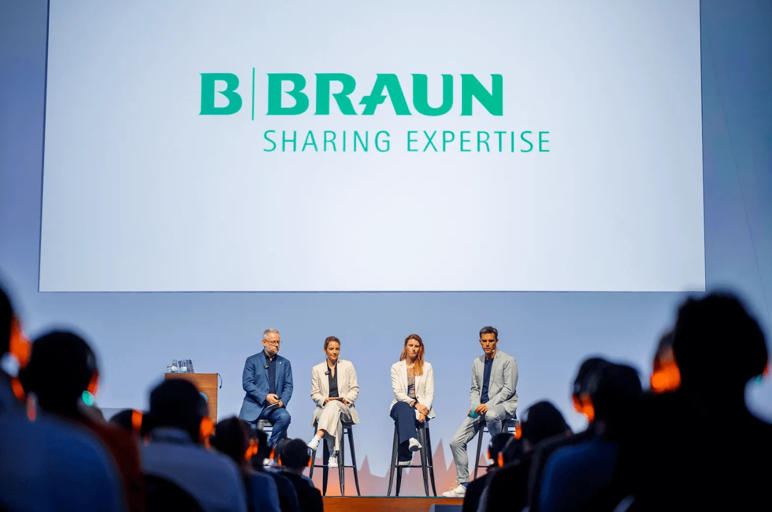 Four panelists sitting on stools in front of a large screen displaying the B Braun logo and 'Sharing Expertise' during a conference.