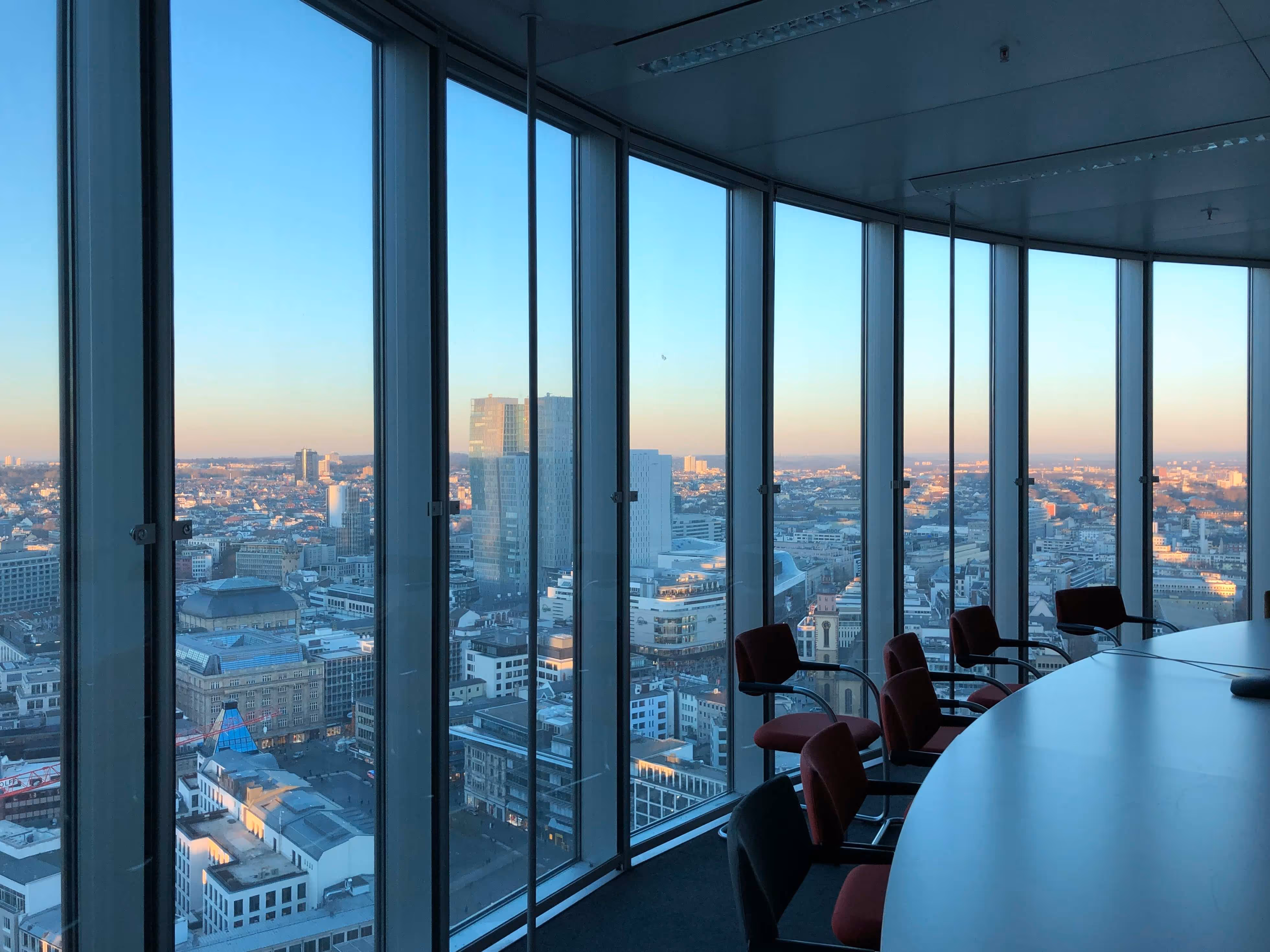 City skyline view from a modern office with curved glass windows, featuring a large table and red chairs in the foreground.