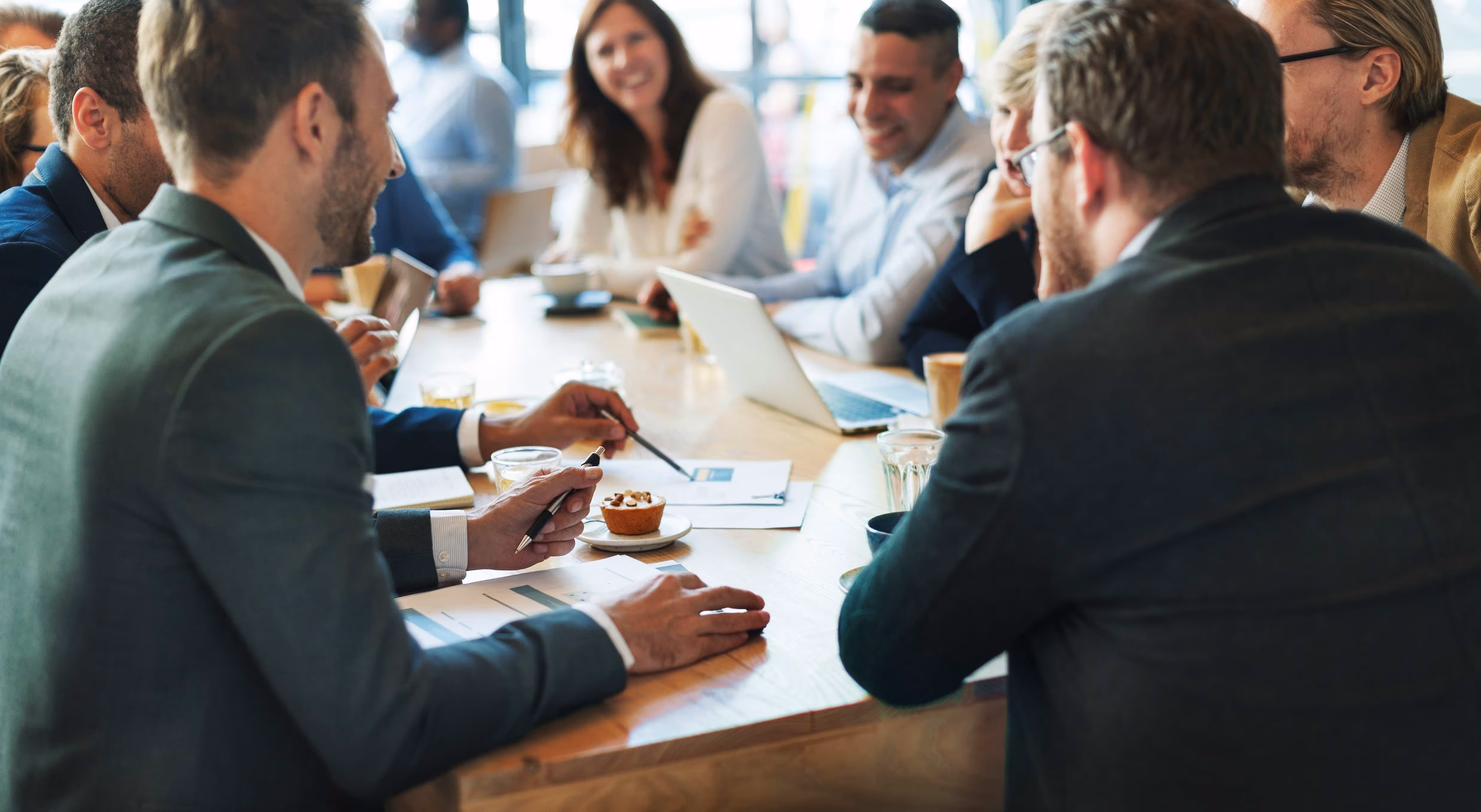 A group of professionals engaged in discussion around a table, with laptops, documents, drinks, and snacks present.