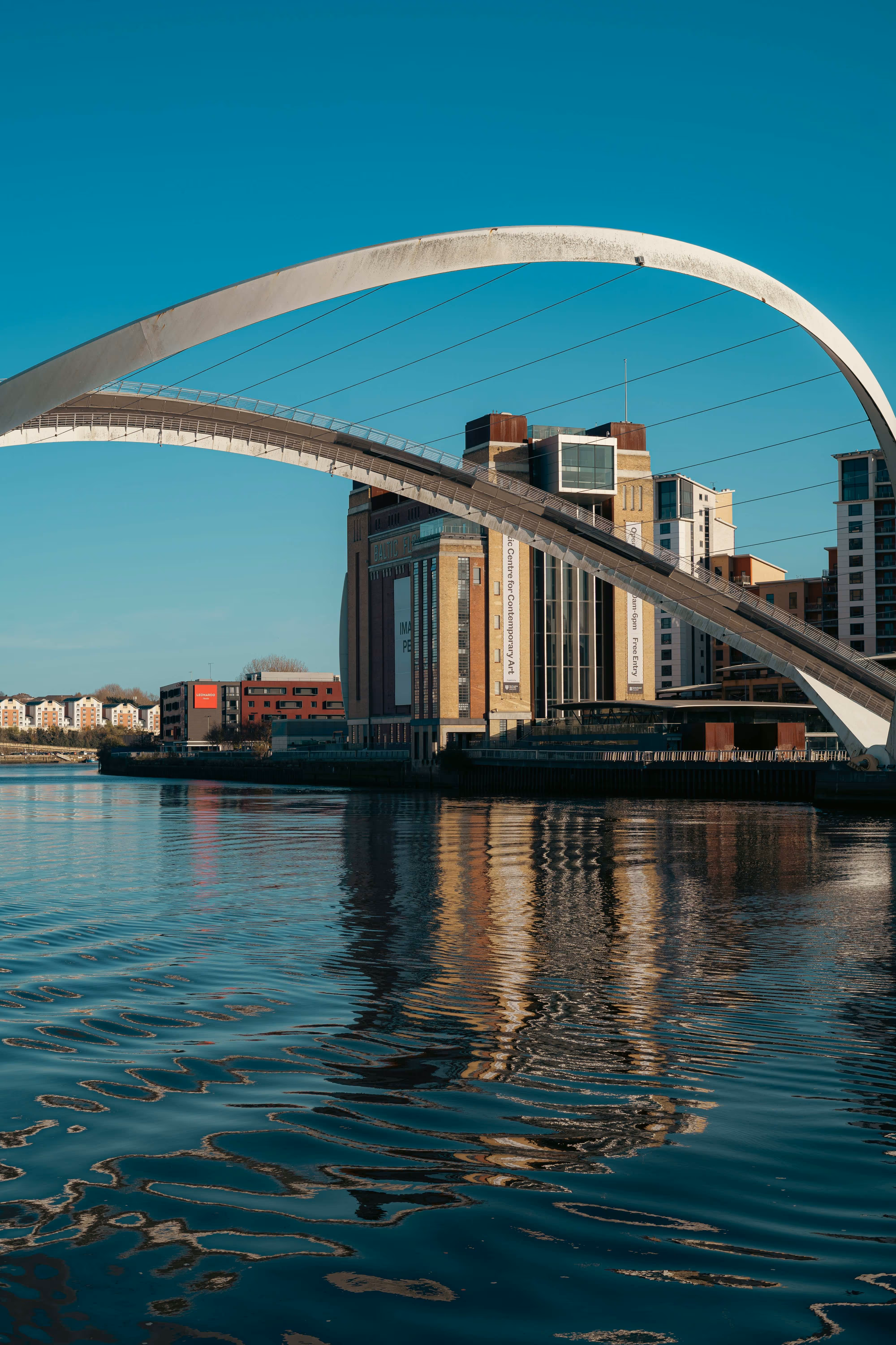 Leeds bridge spans over a river, reflecting nearby buildings and a clear blue sky.