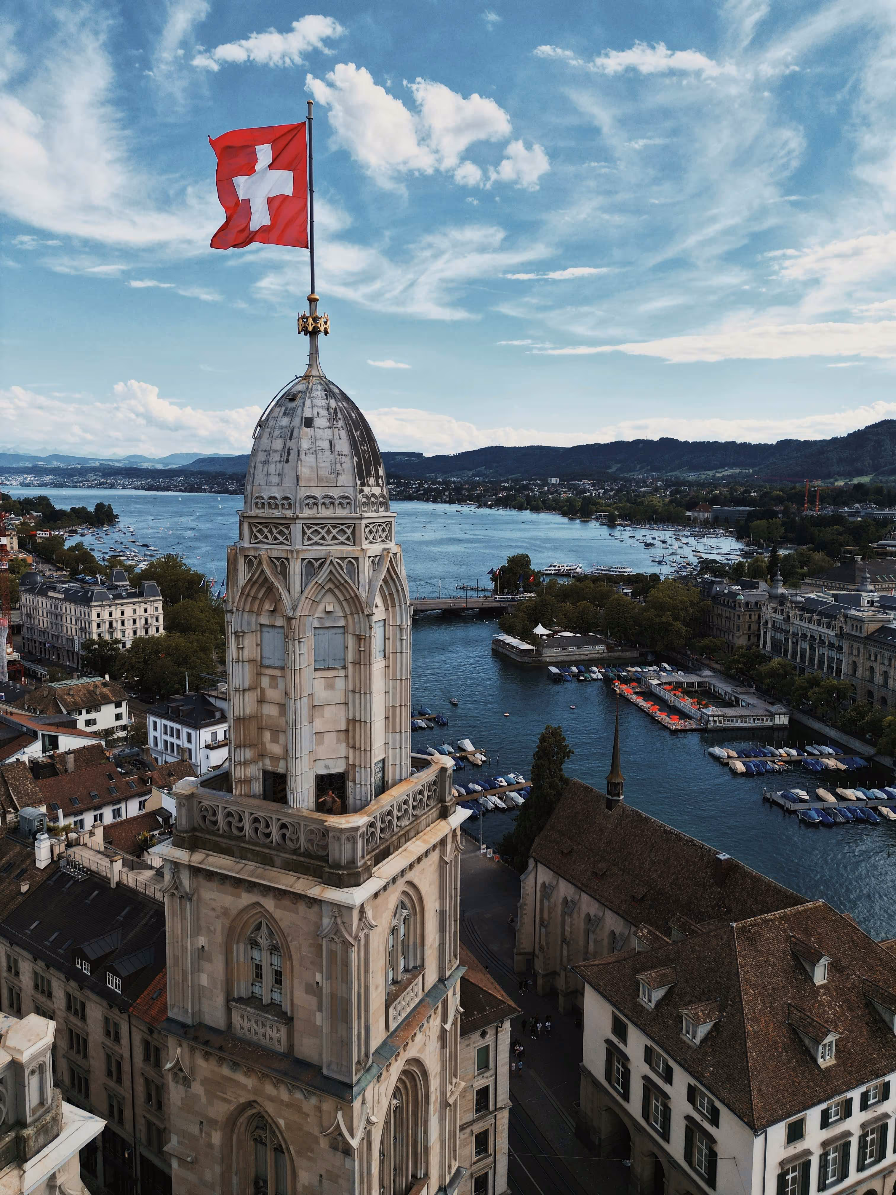 Aerial view of Zurich's skyline featuring a tall tower with a Swiss flag, overlooking the lake and boats under a blue sky.