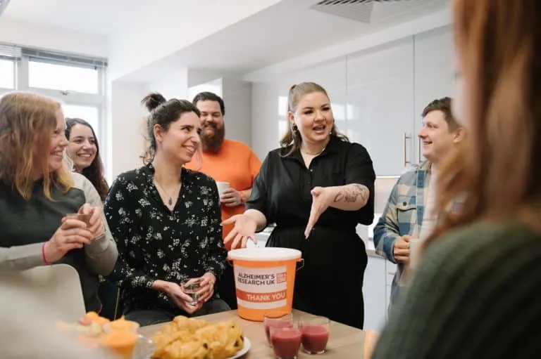 A group of diverse individuals gathers in a kitchen, smiling and engaging, with a donation bucket for Alzheimer's Research UK in front.