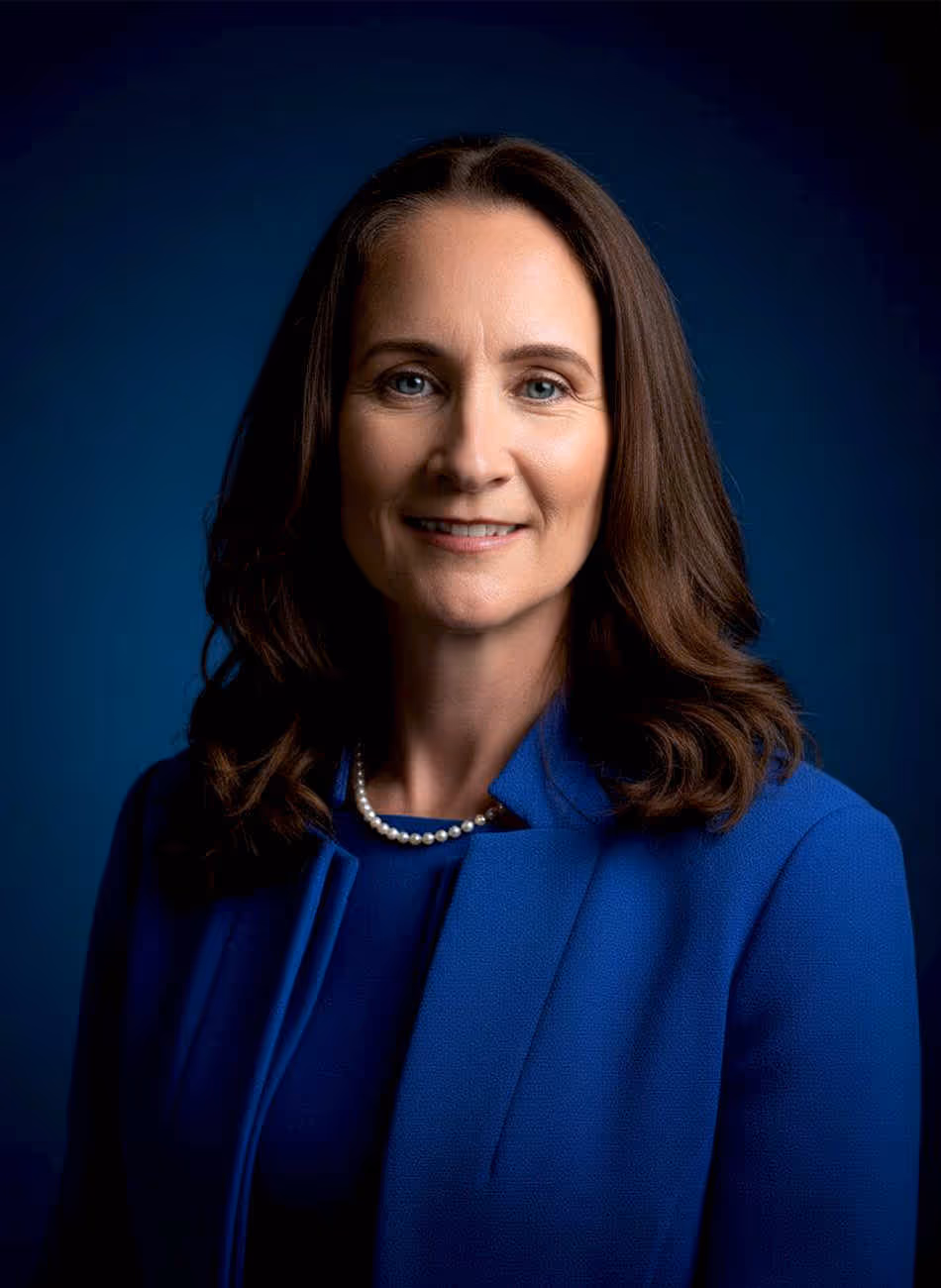 Siobahn Hagerty, lawyer, with long, wavy brown hair, wearing a blue blazer and a pearl necklace, smiles against a dark blue background.