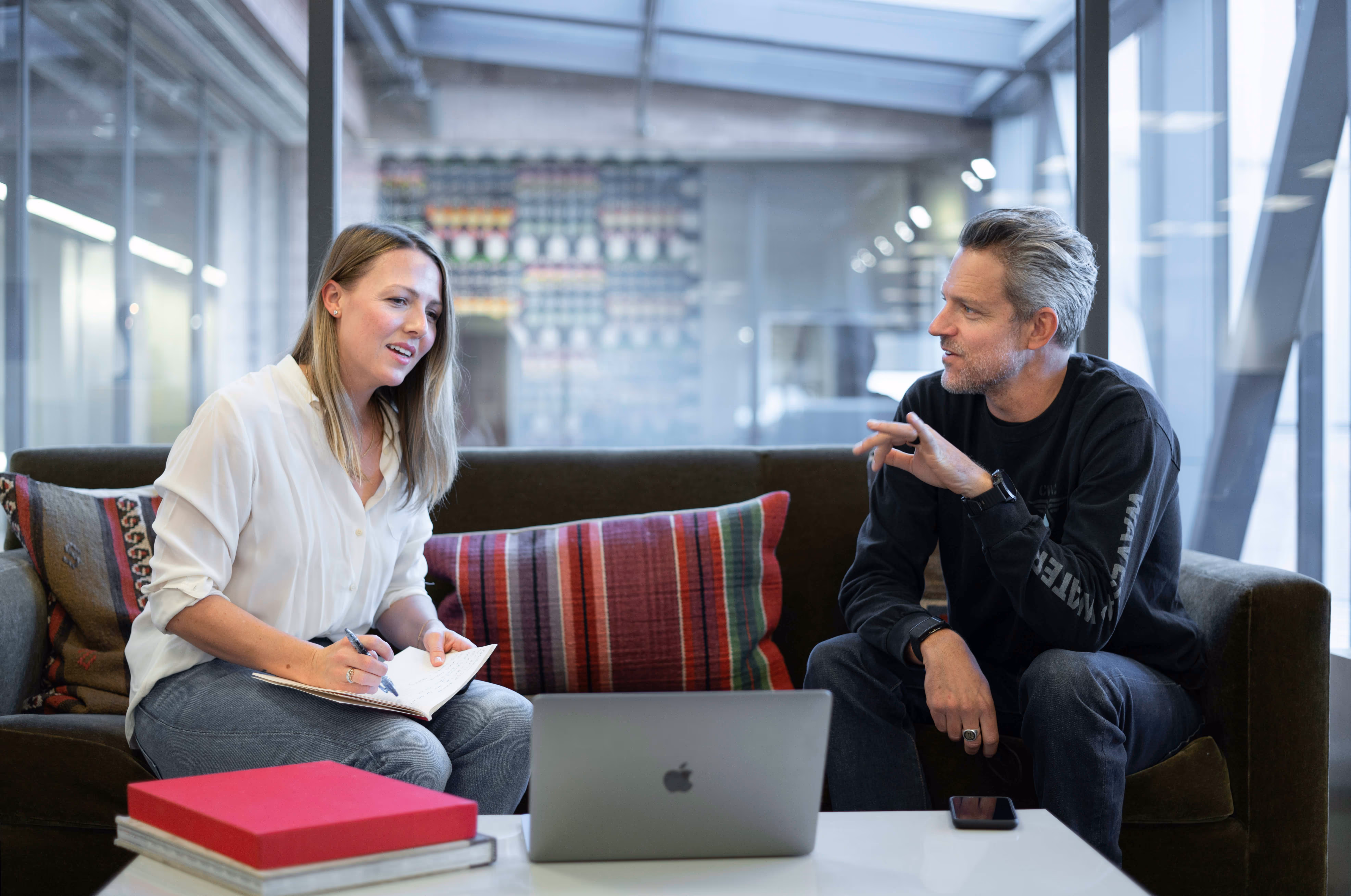 Two professionals engage in a discussion on a sofa, with a laptop and notebook present, in a modern workspace.