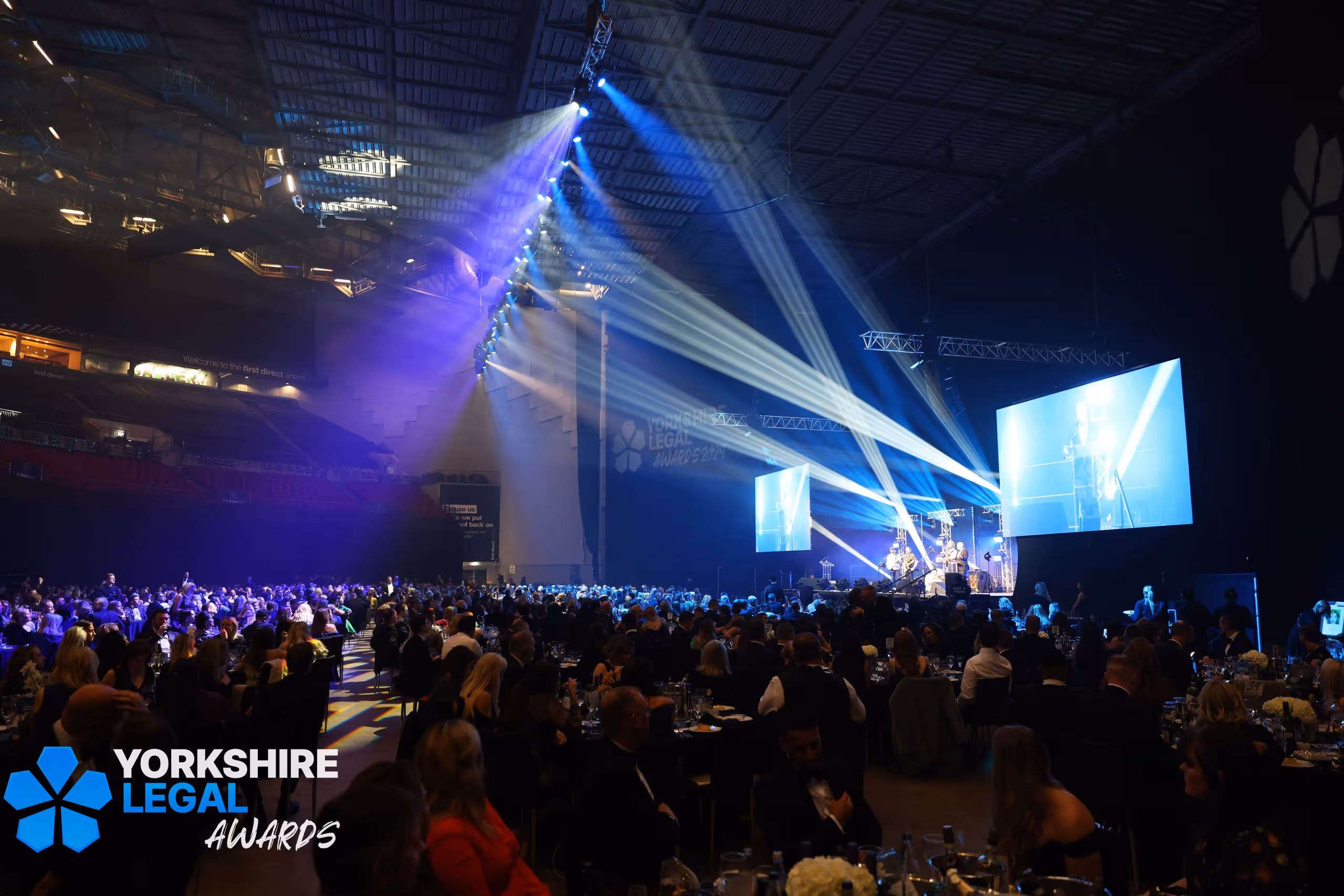 A lively awards ceremony scene with guests seated at tables, stage lights beaming, and a large screen displaying the Yorkshire Legal Awards.