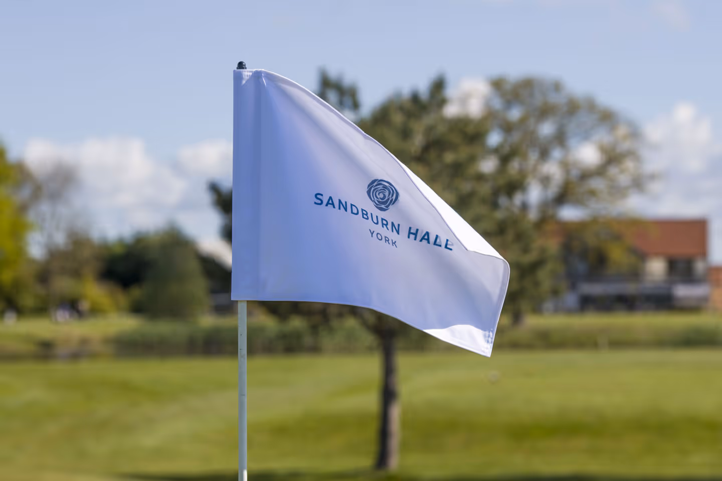 A white flag with the text "SANDBURN HALL YORK" and a logo, standing on a golf course with greenery in the background.