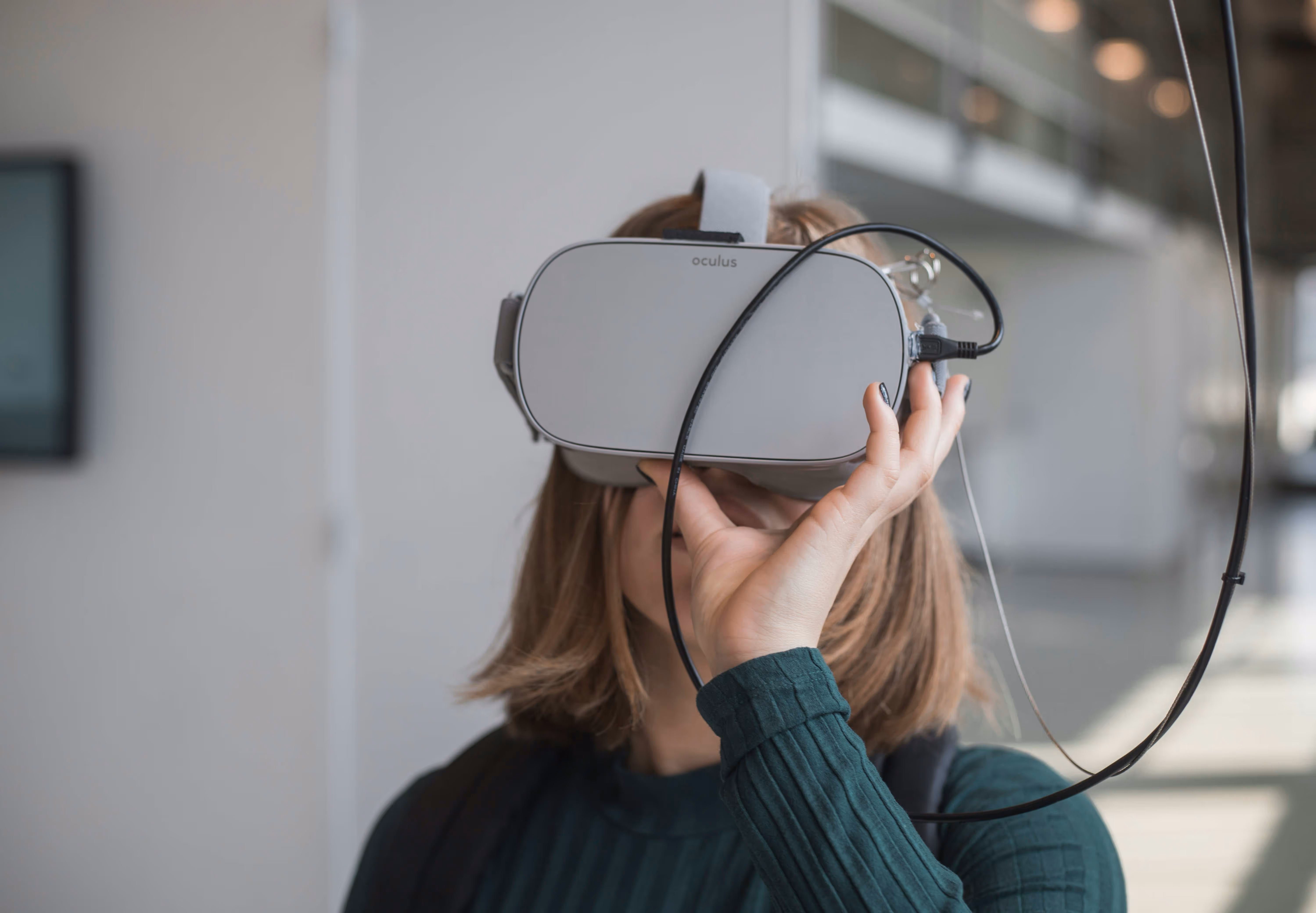 A person wearing a gray Oculus VR headset, holding it with one hand. The background is a blurred indoor space.