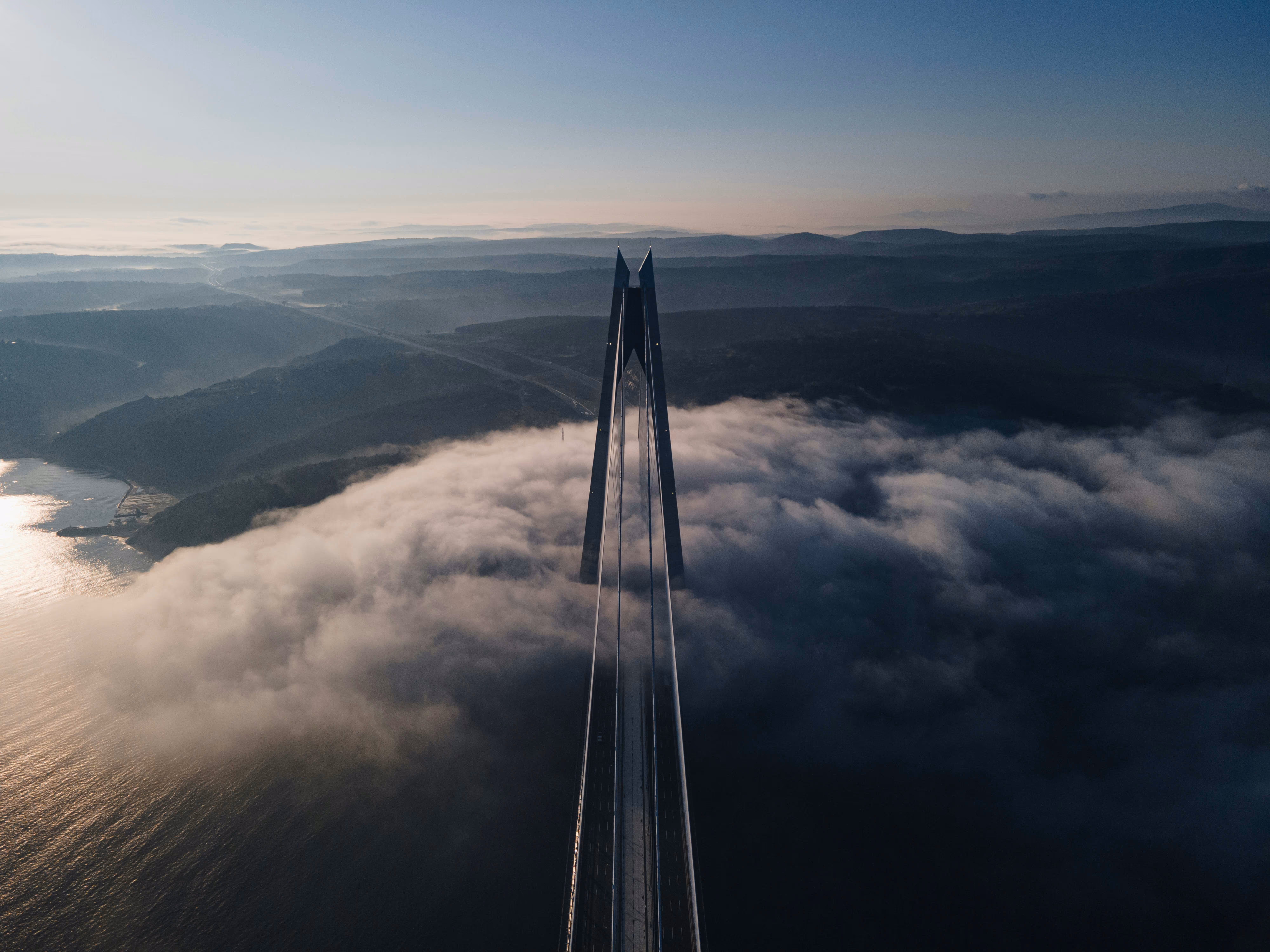 A stunning aerial view of a bridge towering above a sea of fog, with mountains and a river in the background under a clear sky.
